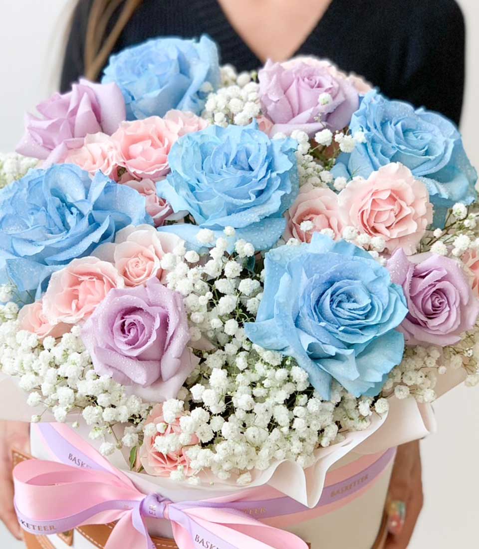 A person in a dark outfit holds a vibrant bouquet featuring pastel blue, pink, and purple roses, surrounded by white baby's breath flowers. The bouquet is wrapped in white and pink paper, tied with a pink ribbon, making it the perfect gift box of vibrant flowers.