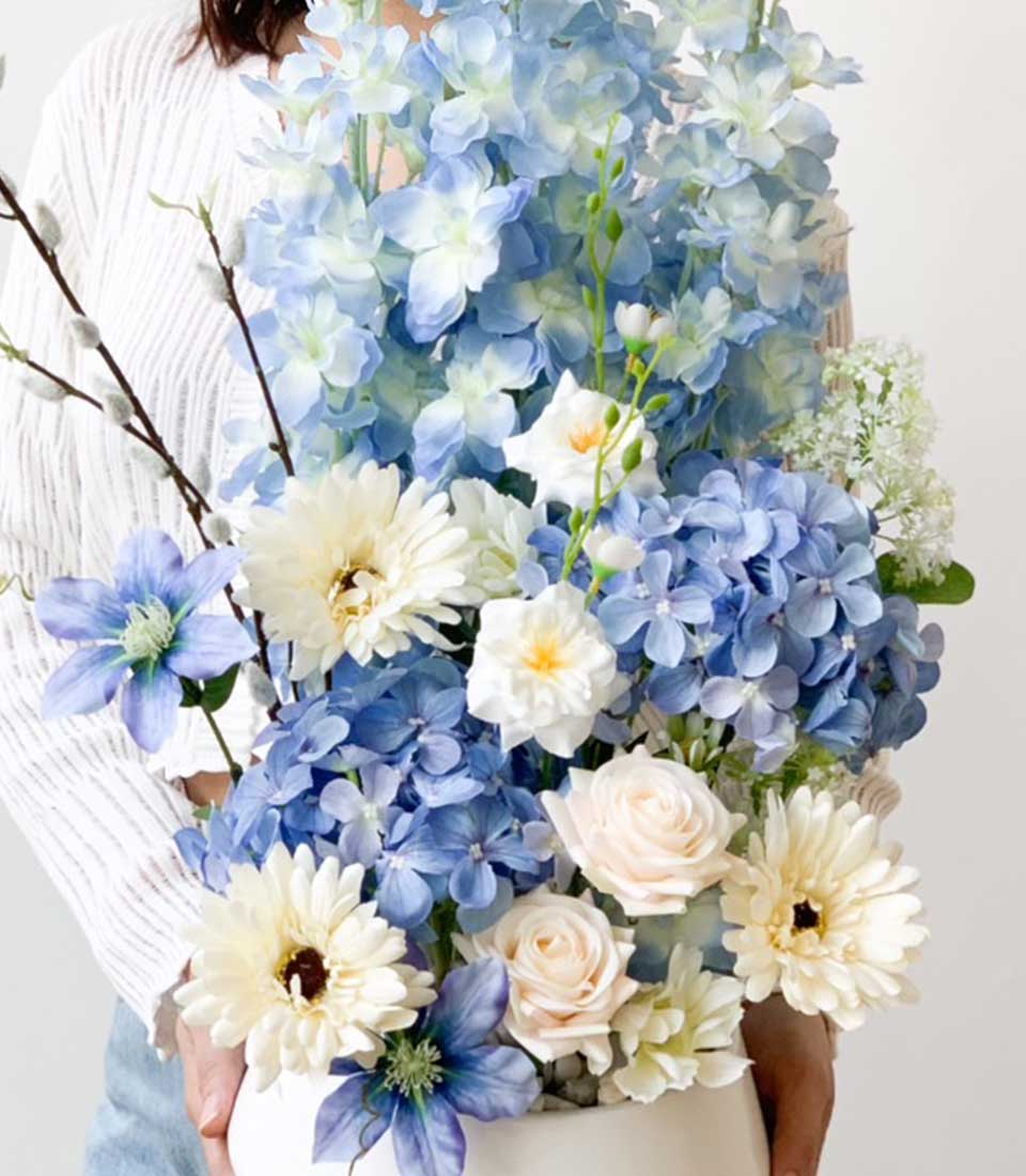 Elegant floral arrangement featuring in white vase a mix of blue and white flowers, including hydrangeas, roses, and daisies, held by a woman in a white top.