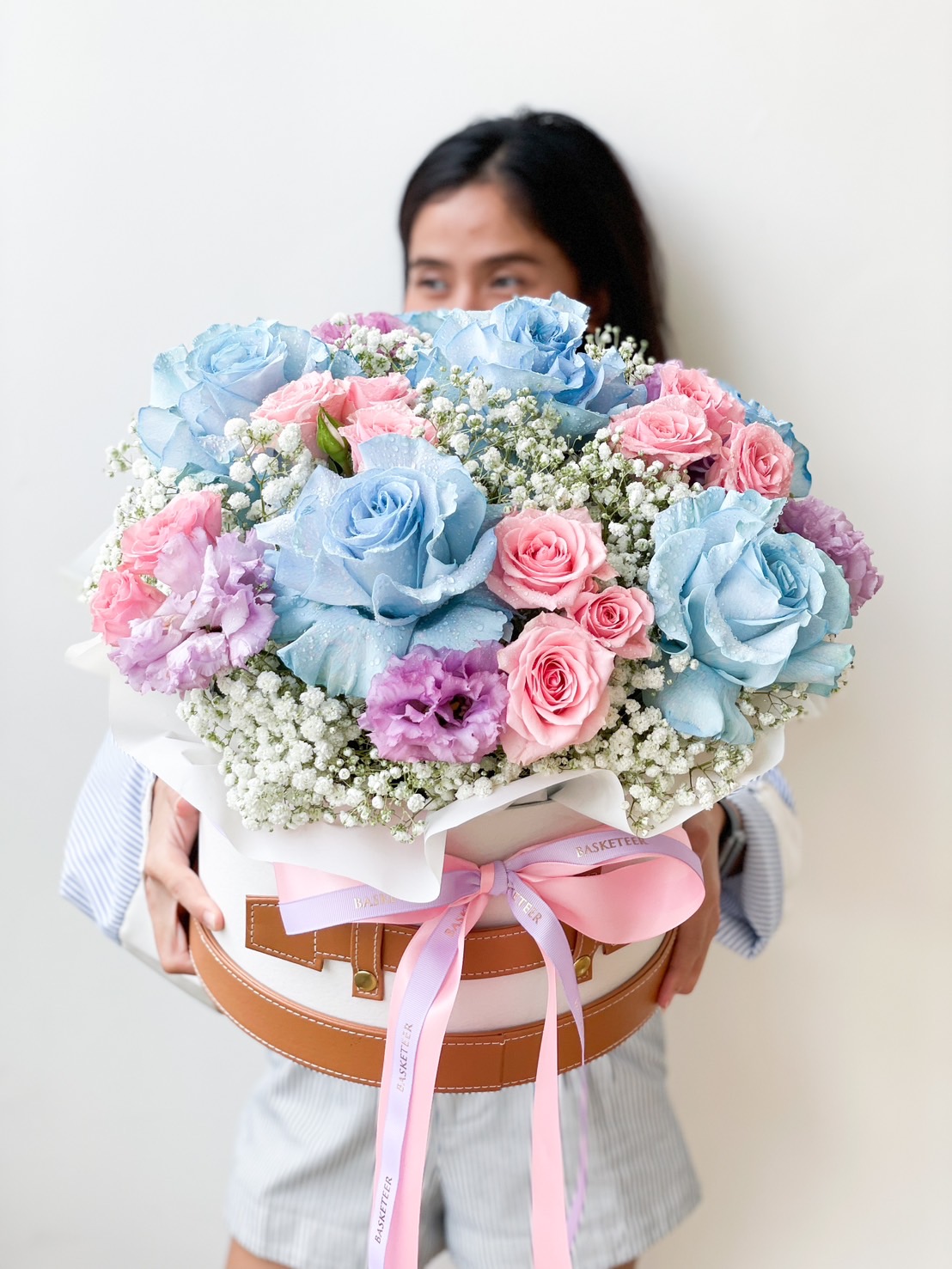 A person holding a large bouquet of vibrant flowers. The bouquet features a mix of blue and pink roses, purple flowers, and white baby's breath, beautifully arranged in a round gift box adorned with a pink ribbon. The background is a plain white wall.
