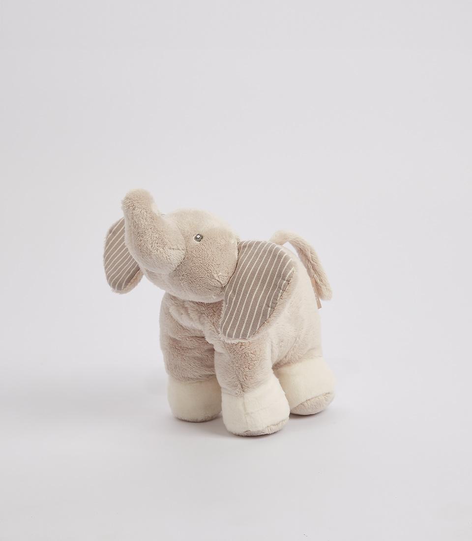 A plush gray elephant toy stands on a white surface. The soft toy has white feet, and its ears are adorned with gray and white stripes. One of its ears sports a small tag. The plush gray elephant has an upward-curved trunk and a friendly expression.