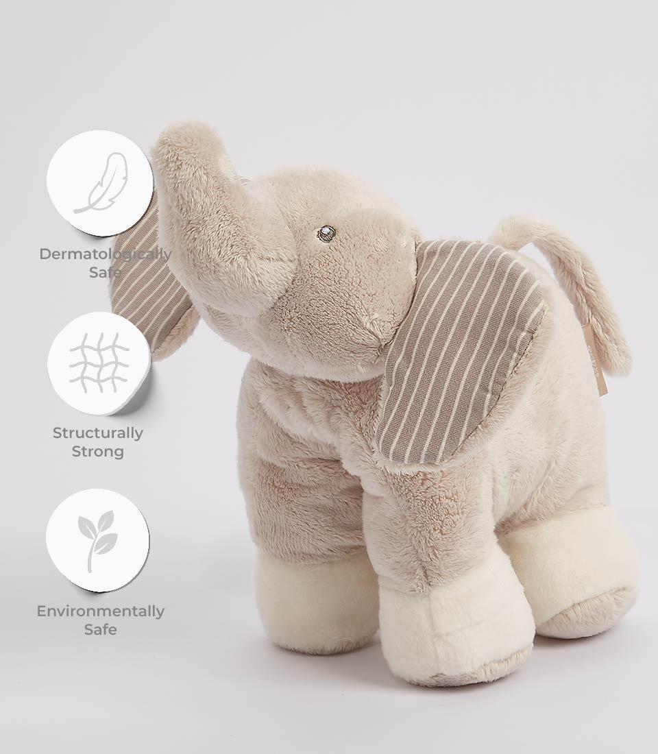 A plush gray elephant toy stands on a white surface. The soft toy has white feet, and its ears are adorned with gray and white stripes. One of its ears sports a small tag. The plush gray elephant has an upward-curved trunk and a friendly expression. Next to the soft toy are three circular icons with text: 