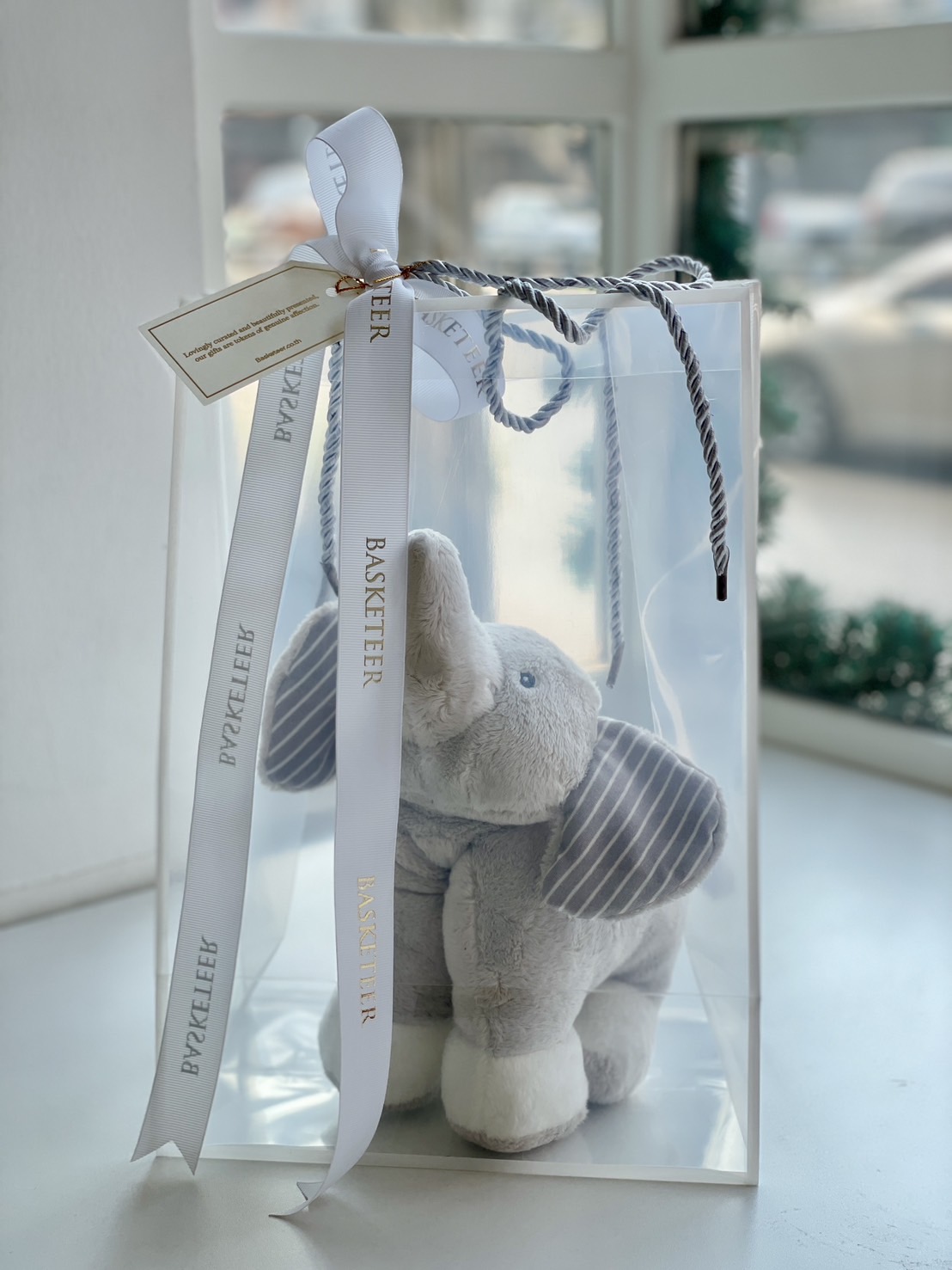 A soft grey elephant plush toy with striped ears, beautifully packed in a luxury transparent gift bag with a white Basketeer ribbon.