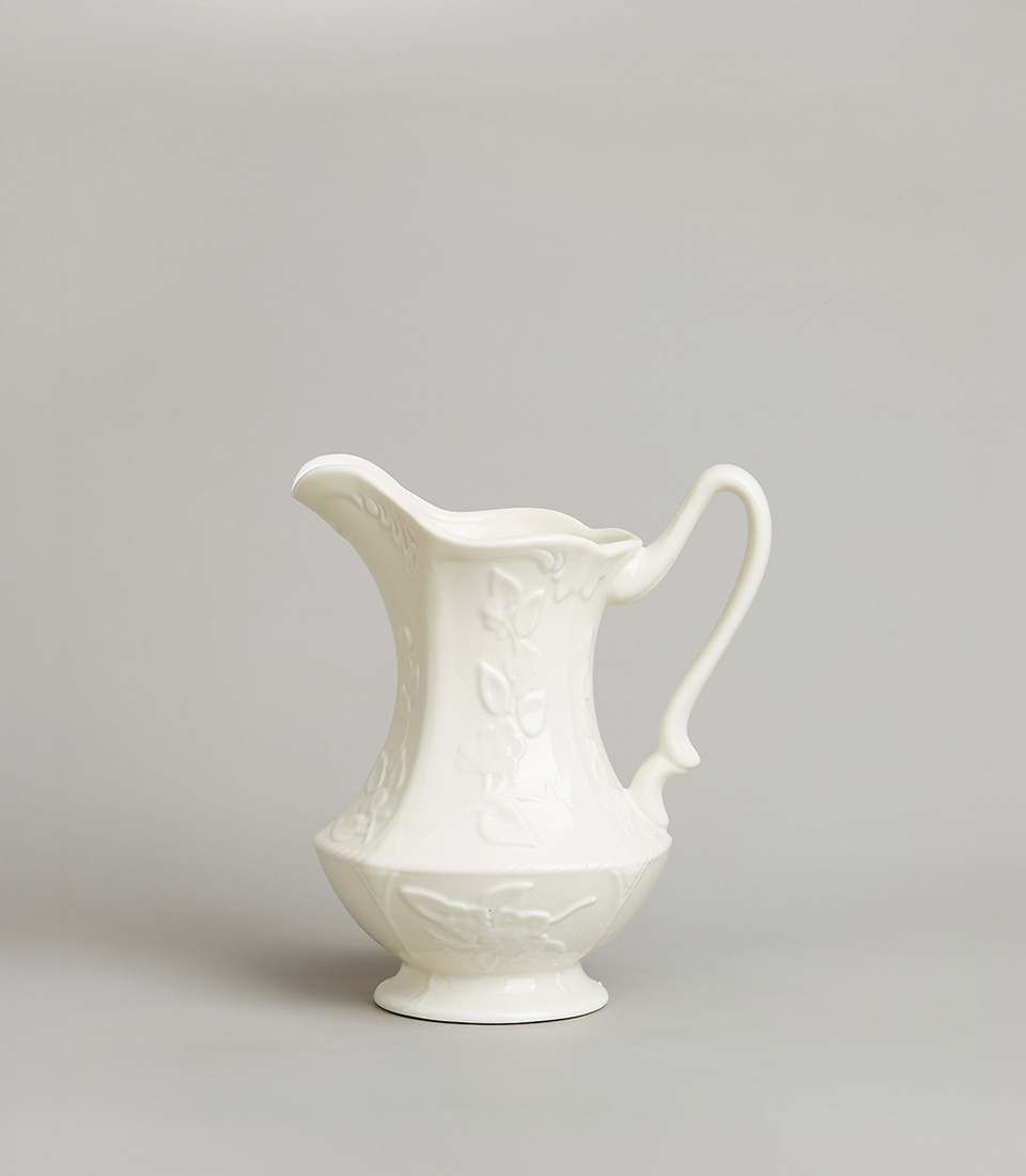 Elegant white ceramic pitcher with an embossed floral design, featuring a gracefully curved handle and spout, on a light grey background.