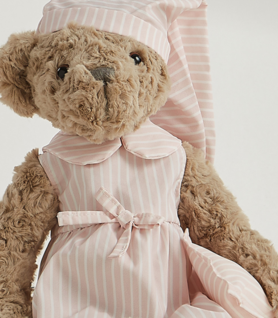A fuzzy gray teddy bear in a white and pale pink striped dress and matching sleep cap stands upright on a plain white background, epitomizing the charm of soft toys.