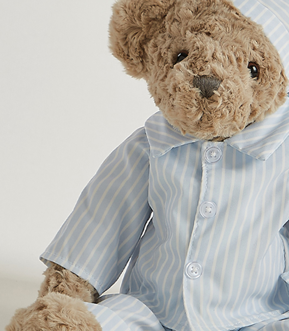 A plush teddy bear in striped pajamas and a matching nightcap stands against a plain white background. The soft toy's fur is grey, and it has a slightly serious expression on its face.