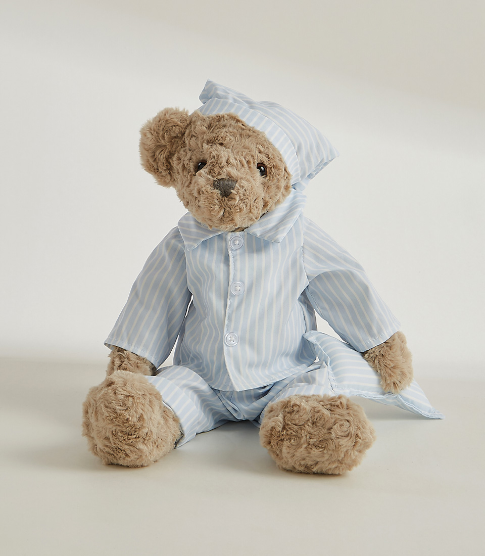 A plush teddy bear in striped pajamas and a matching nightcap stands against a plain white background. The soft toy's fur is grey, and it has a slightly serious expression on its face.