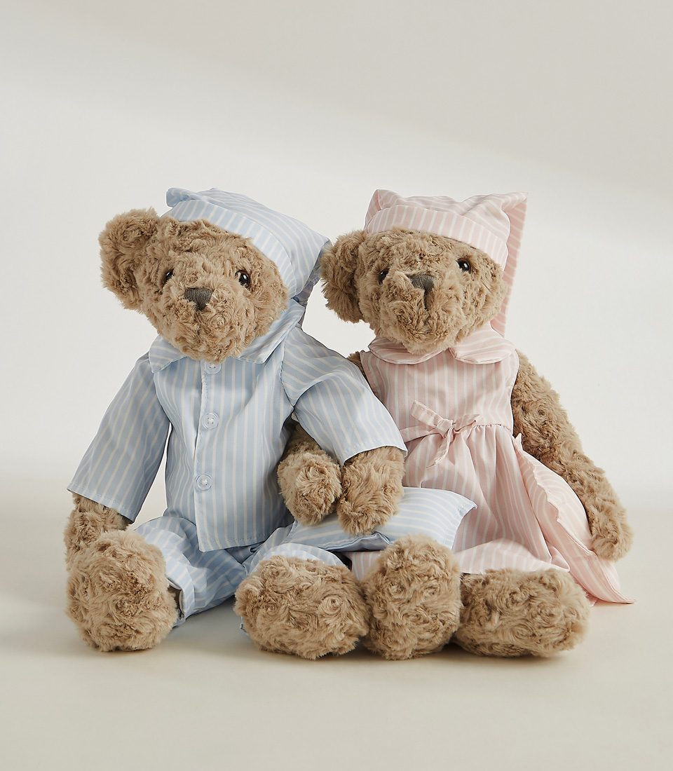 The Sleepytime Teddy Bear Duo Soft Toys feature two plush teddy bears dressed in striped pajamas and nightcaps. The larger bear wears a pink nightgown with a matching pink cap, while the smaller bear is snug in a blue pajama set with a matching blue nightcap.