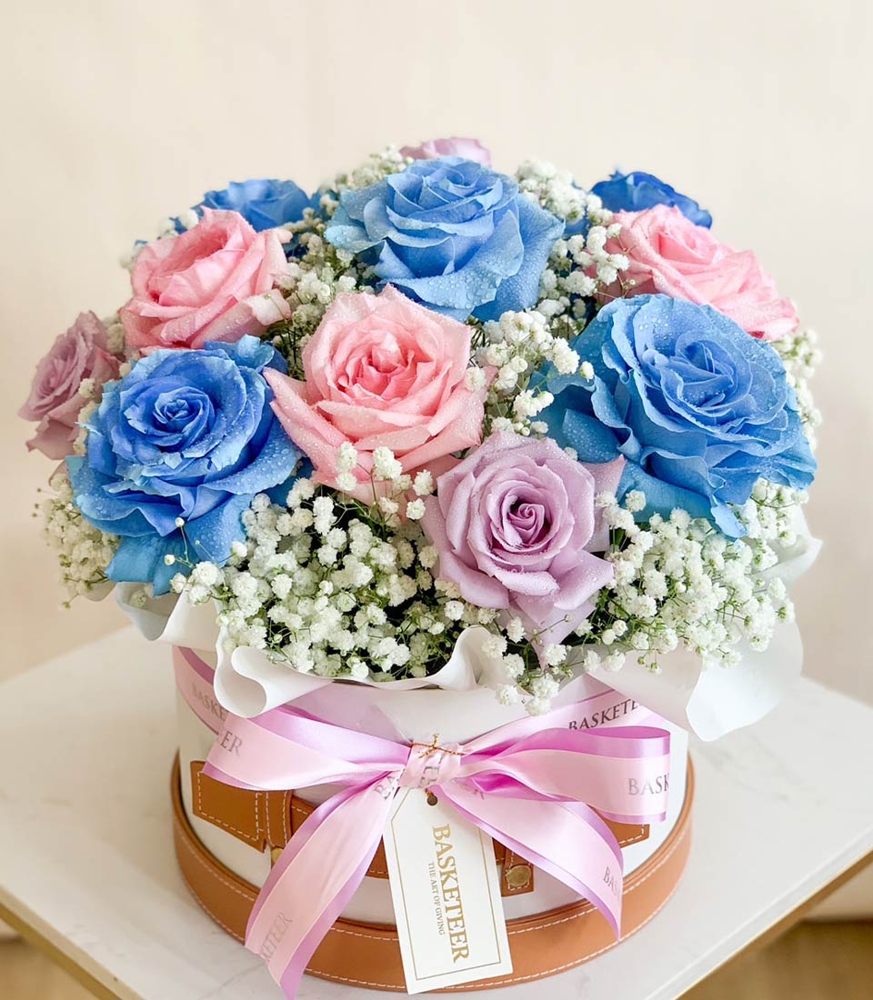 An elegant arrangement of blue, pink, and lavender roses with baby’s breath, beautifully presented in a white gift box with pink ribbons.