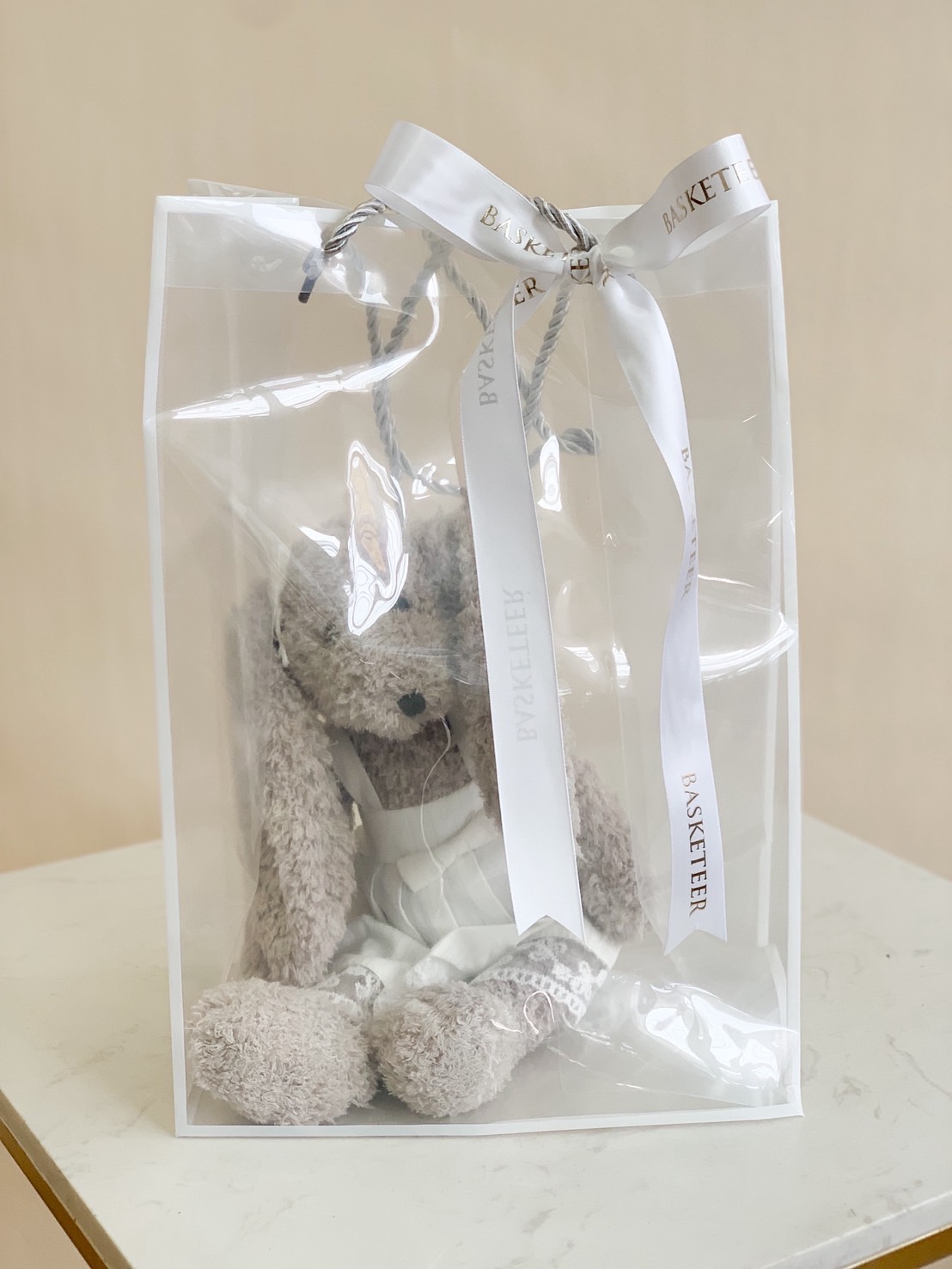 Soft grey plush bunny wearing a white outfit, seated inside a clear premium gift bag with white Basketeer ribbon.