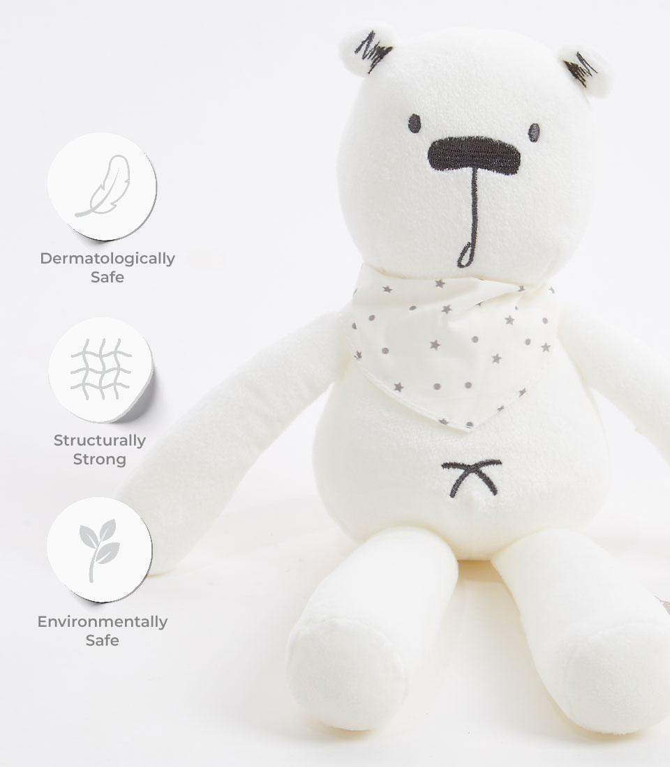 A cuddly white teddy bear with simple black facial features, wearing a white scarf adorned with small star patterns. Surrounding icons highlight that this soft toy is dermatologically safe, structurally strong, and environmentally friendly.