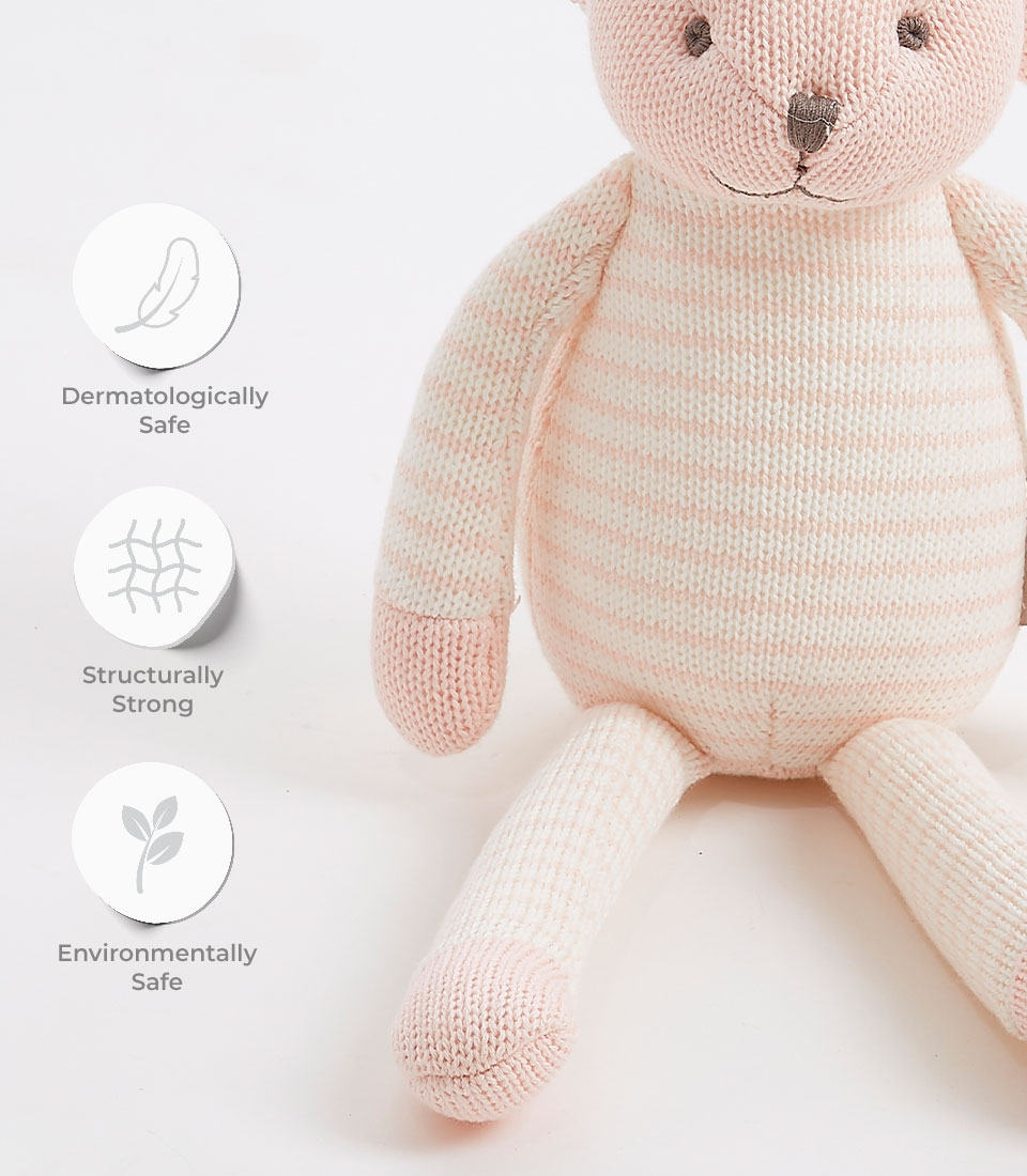 A cuddly, striped pink and white stuffed animal bear sits next to icons and text. The icons indicate the stuffed animal is dermatologically safe, structurally strong, and environmentally safe. The bear has stitched eyes and nose, and is positioned on a white background.