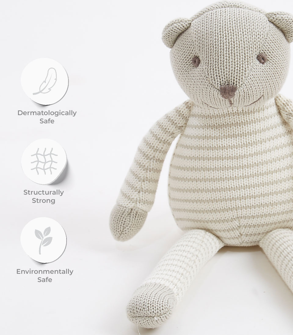 A gray striped knitted teddy bear with beige and white accents is displayed. Three icons with text indicate the soft toy's features: 