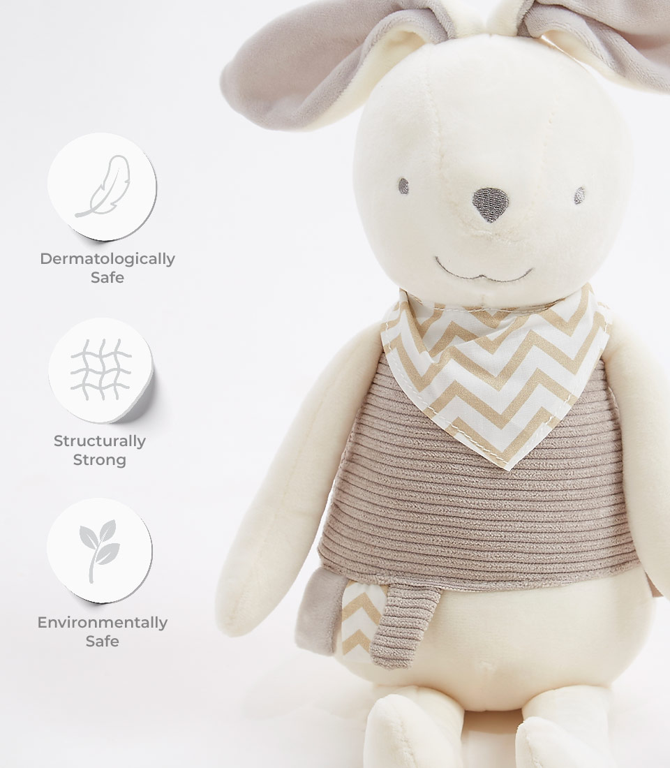 A gray bunny plush toy with floppy ears, a chevron-patterned bandana, and a striped body. Three icons with text to the left state: Dermatologically Safe, Structurally Strong, and Environmentally Safe. This soft toy is set against a white background.
