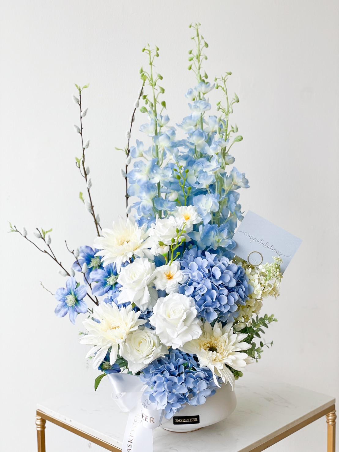 A stunning blue floral arrangement in a white vase boasts a variety of blue and white flowers, including roses, hydrangeas, dahlias, and delphiniums. Nestled among the blooms is a greeting card with 