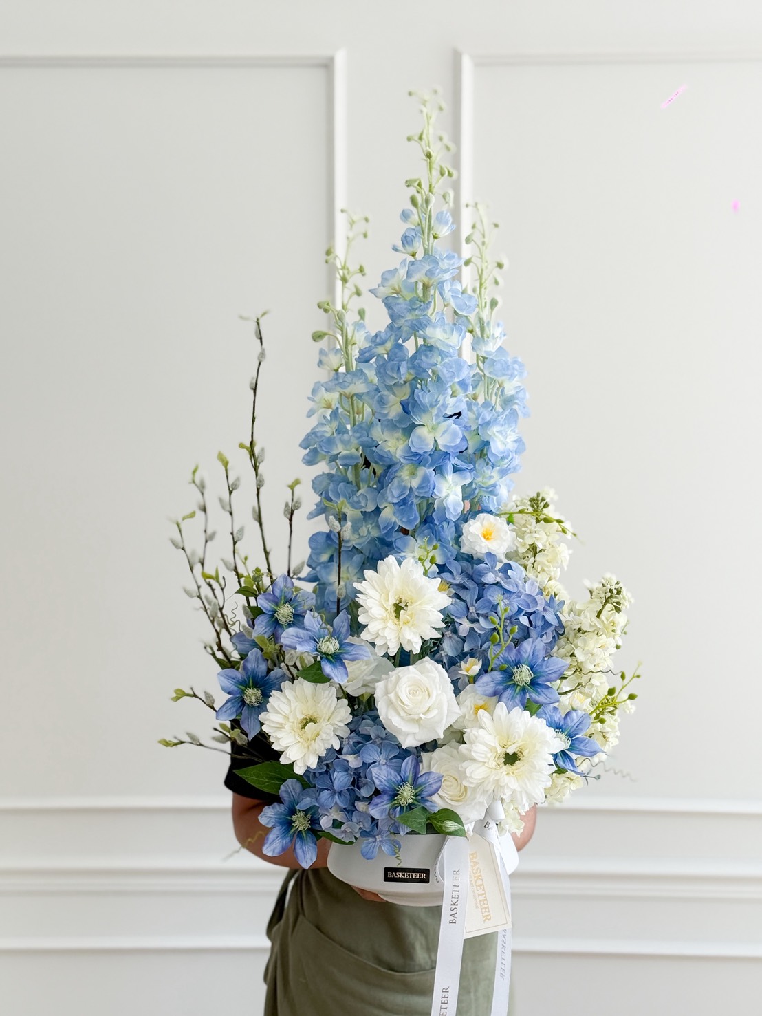 A person holds a stunning blue floral arrangement featuring blue delphiniums, white roses, and white chrysanthemums with greenery, set against a white, minimal background.