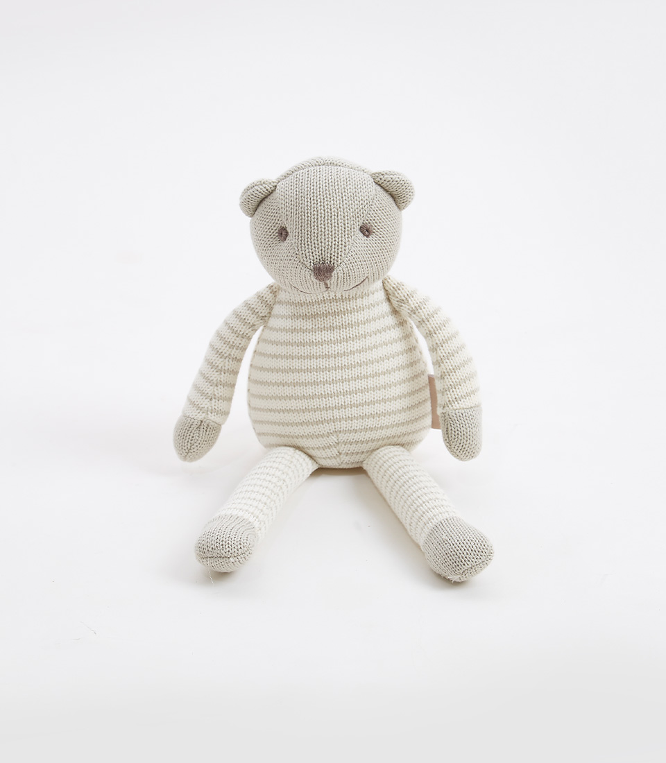 A knitted teddy bear with a gray striped pattern in shades of beige and white sits upright against a plain white background. The soft toy has a round head with small ears, a stitched smile, and simple eyes. Its limbs are long and floppy, matching the striped body.