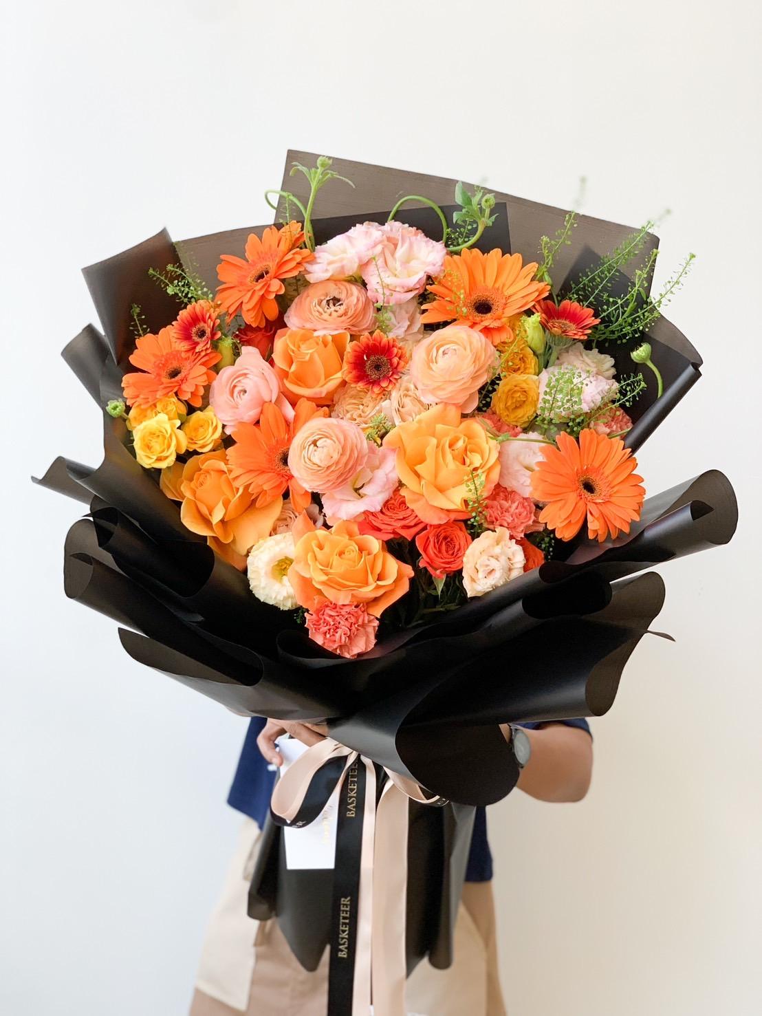A person holding a large Sunshine Splendor Bouquet wrapped in black paper. The bouquet features a variety of colorful flowers, including orange roses, orange gerbera daisies, pink roses, and other small orange and pink blossoms against a plain white background.