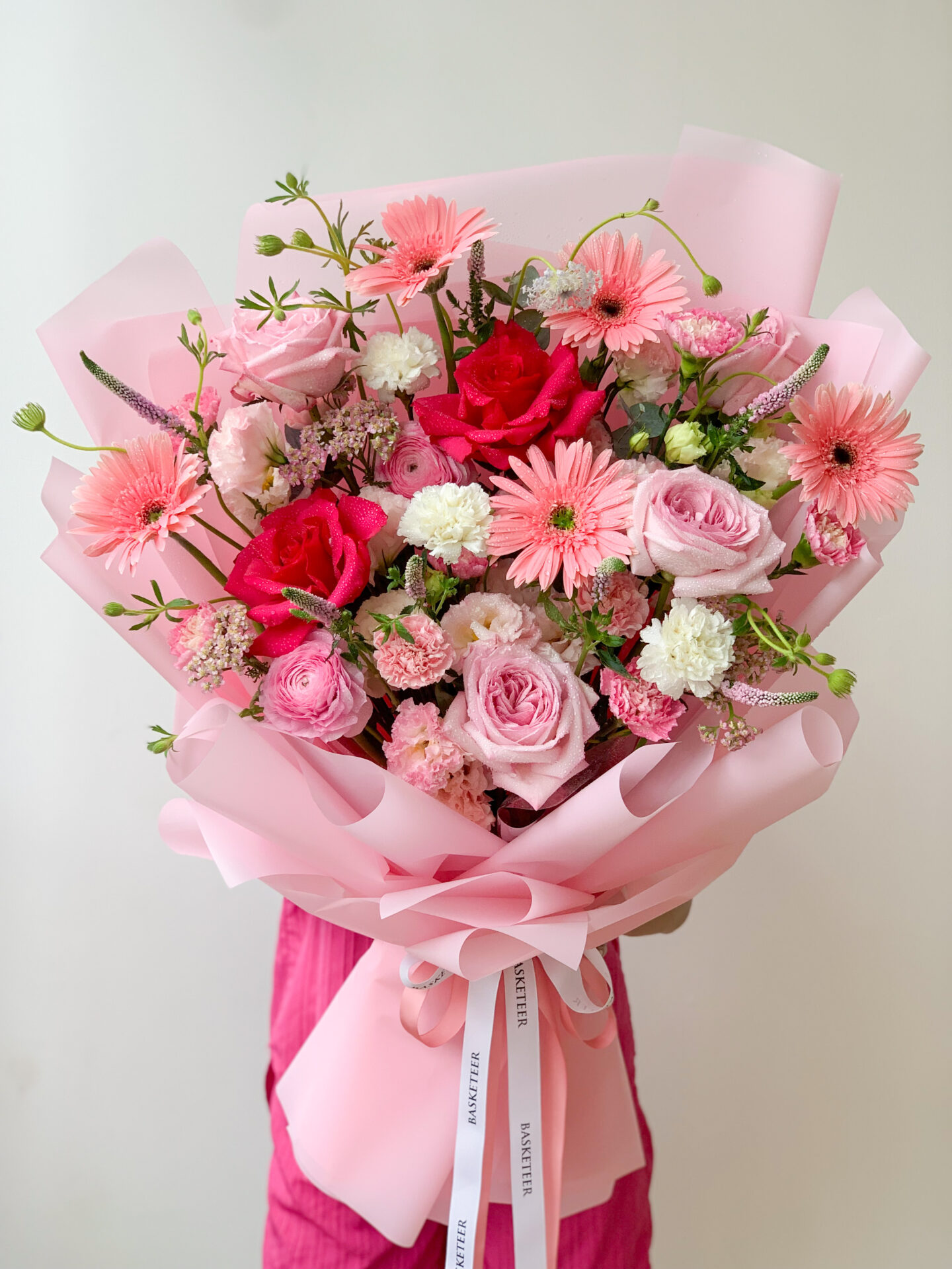 A delightful Pink Blossom Bouquet featuring a vibrant mix of pink and white roses, gerbera daisies, and carnations, wrapped in pink paper with charming ribbon accents. The bouquet is held against a plain white background.