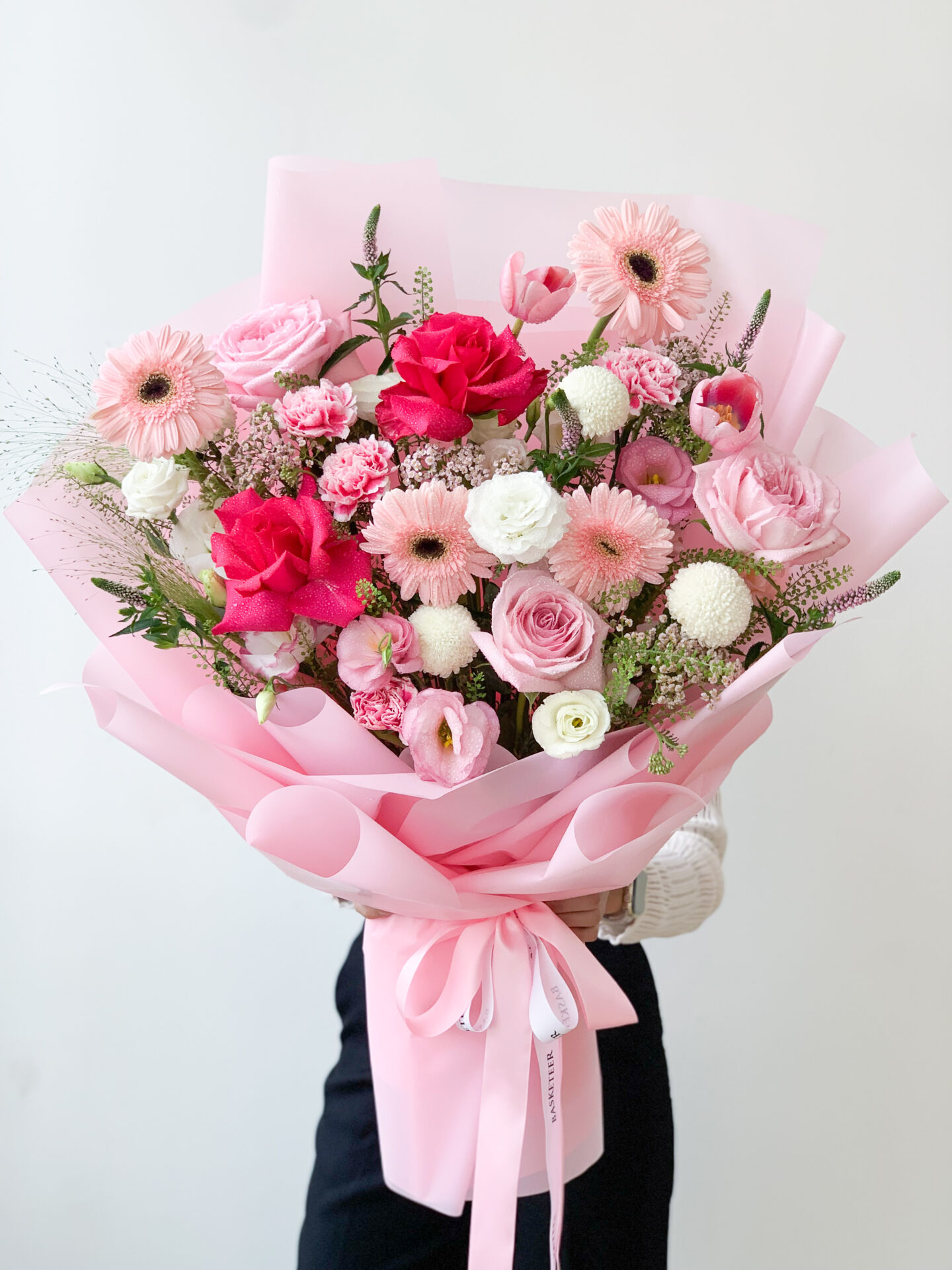 A person holding a delightful pink blossom bouquet wrapped in pink paper. The bouquet features a variety of flowers, including pink roses, gerberas, white chrysanthemums, and pink lisianthus, arranged with greenery. The person is wearing a white sweater and black pants.
