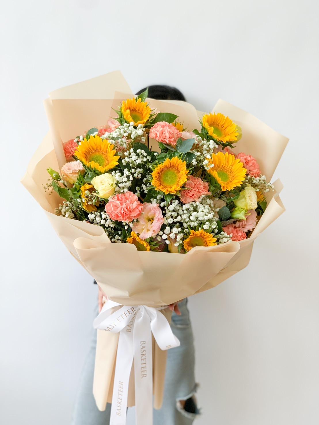 A person holding a large Sunny Delight Bouquet, including sunflowers, pink carnations, white baby's breath, and greenery, wrapped in light beige paper with a white ribbon at the bottom. The person is mostly obscured by the bouquet.