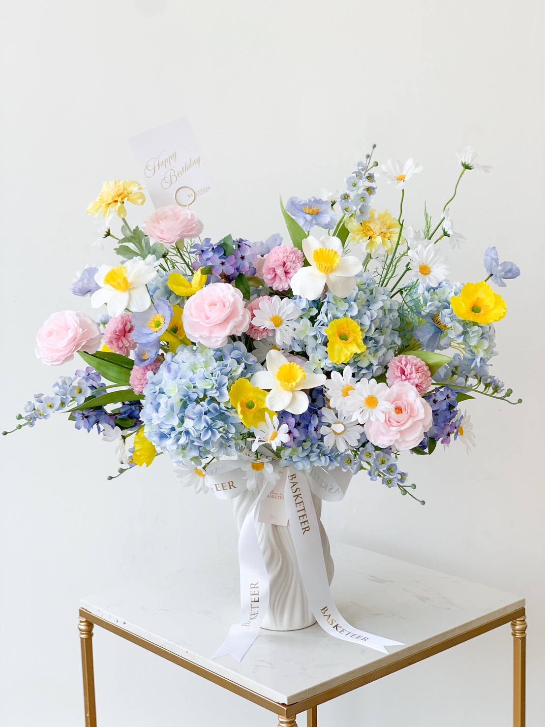 A large, vibrant floral arrangement sits in a white ceramic vase on a small table. This energetic spring bloom features pink roses, yellow and white daisies, blue hydrangeas, and purple delphiniums. A small card reading 