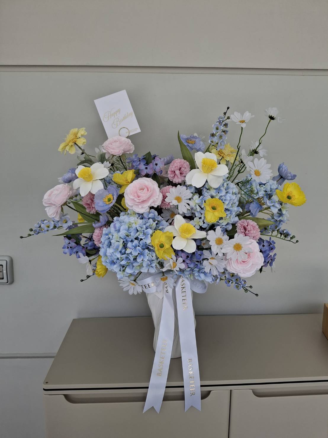 A large, elegant bouquet of flowers featuring pink roses, blue hydrangeas, white daisies, and yellow poppies is arranged in an artificial flower vase. A card is attached reading 