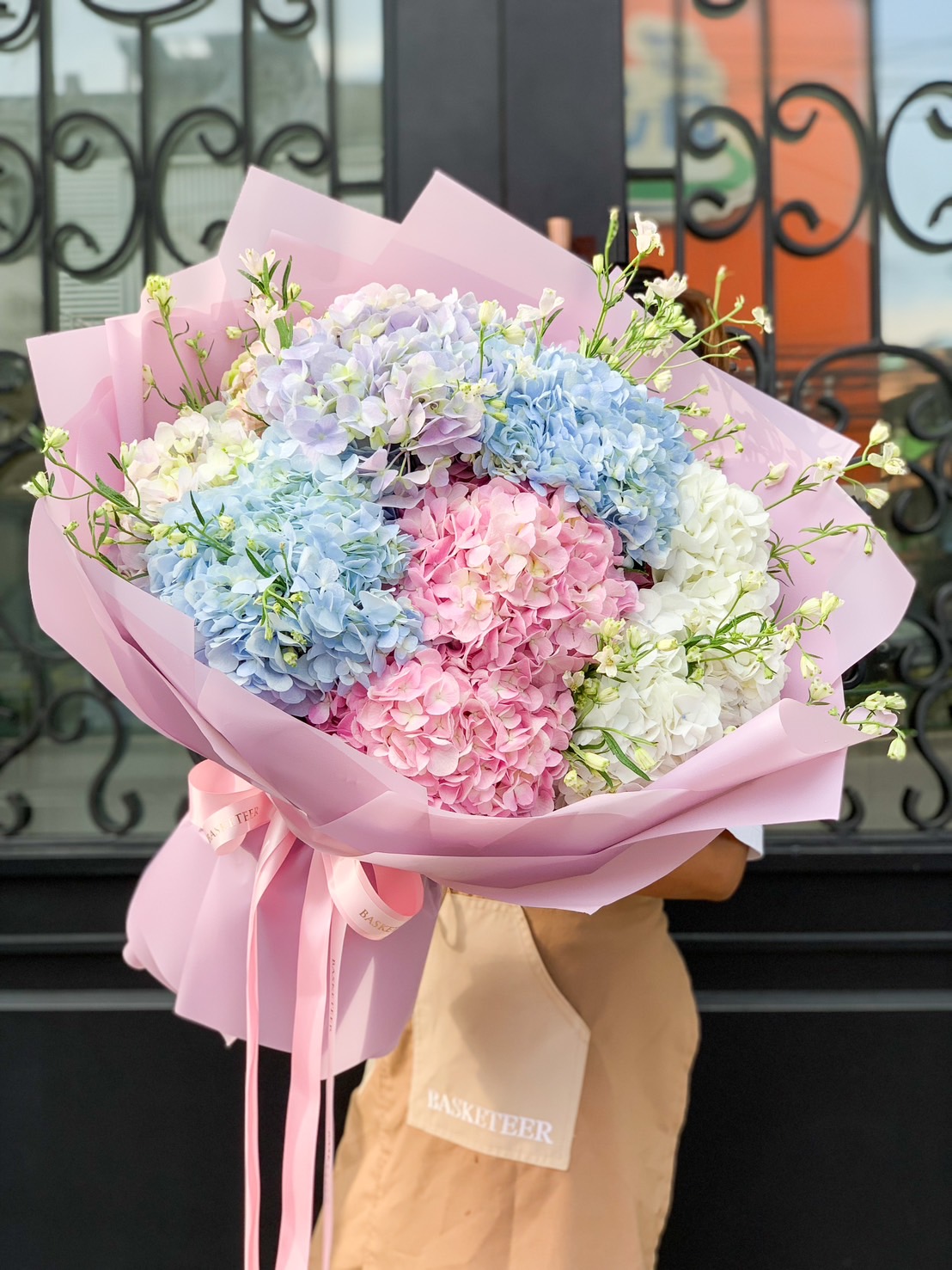 A person holds a large bouquet of hydrangeas wrapped in light pink paper and tied with pink ribbons. This Hydrangea Symphony features blooms in shades of blue, pink, and white, complemented by small white flowers. A metal gate with intricate designs is visible in the background.
