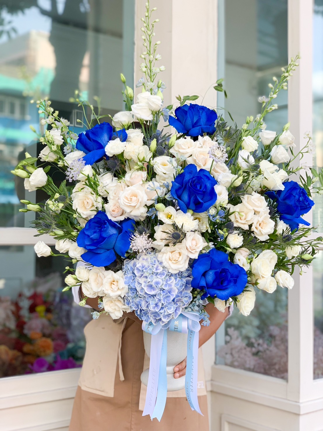 A person holds a large, elegant blue and white floral arrangement in front of a flower shop, obscuring their face. The bouquet features vibrant blue roses surrounded by white roses, delicate white flowers, and greenery. Adorned with light blue hydrangea and ribbons, it captures pure elegance.