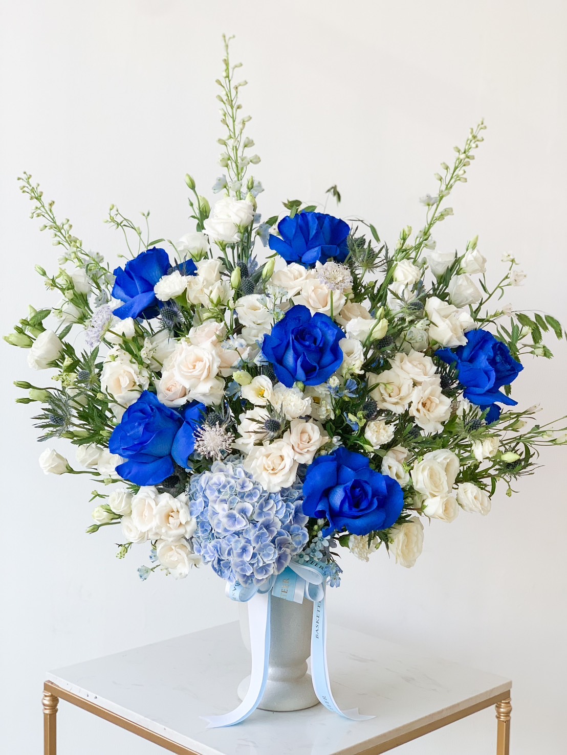 An elegant blue and white floral arrangement in a white vase, featuring vibrant blue roses, white roses, and lush greenery. A small blue hydrangea cluster stands out in front, complemented by a light blue ribbon adorning the vase. The background remains plain to emphasize its beauty.