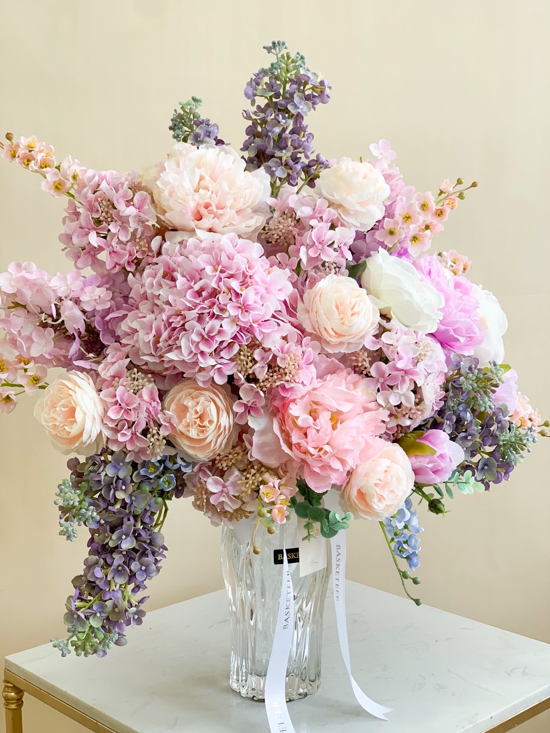 Elegant pastel flower arrangement with pink peonies, blush roses, and purple hydrangeas in a clear glass vase.