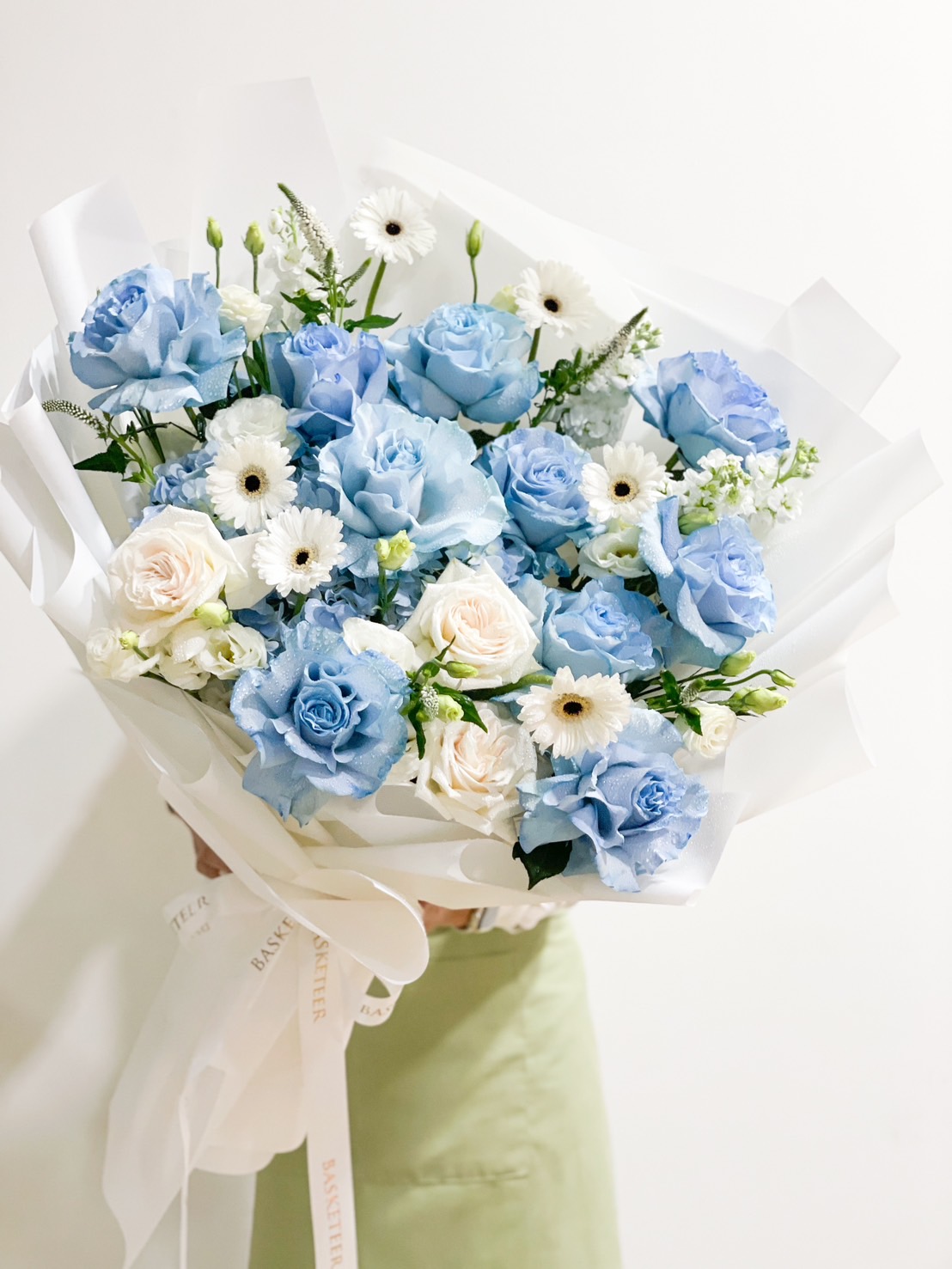 The Serene Blue Bliss Flower Bouquet, with its detailed and abundant mix of blue roses, white roses, white daisies, and other small white flowers, is elegantly wrapped in white paper. This serene arrangement is held against a plain white background.