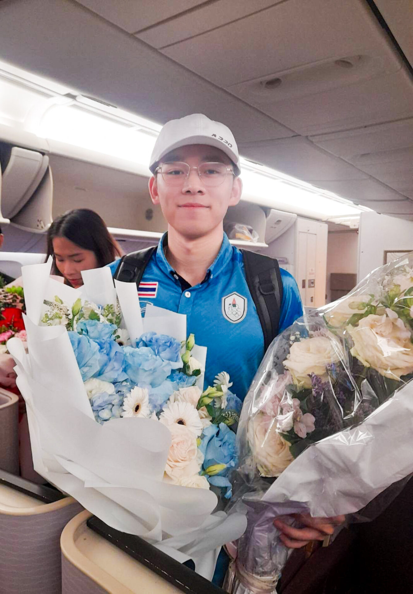 A person wearing a blue shirt and a white cap is holding two large bouquets of flowers, including a Serene Blue Bliss Flower Bouquet wrapped in white paper with blue and white blossoms. They are standing inside an airplane cabin, with another person visible in the background.