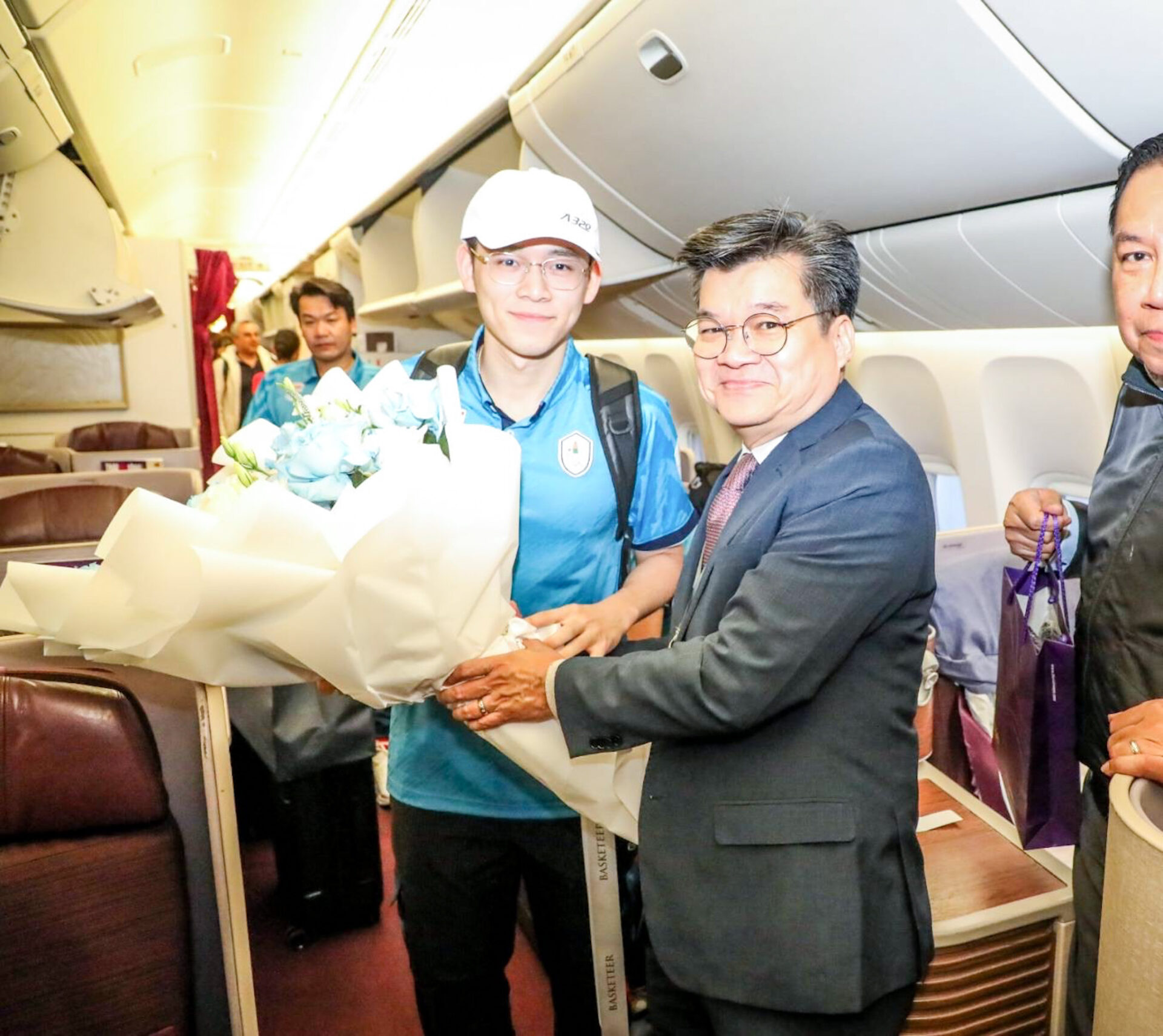 A young man in a serene blue shirt and white cap smiles as he receives a blissful flower bouquet from a man in a suit and tie. They are inside an airplane, and other passengers can be seen in the background.