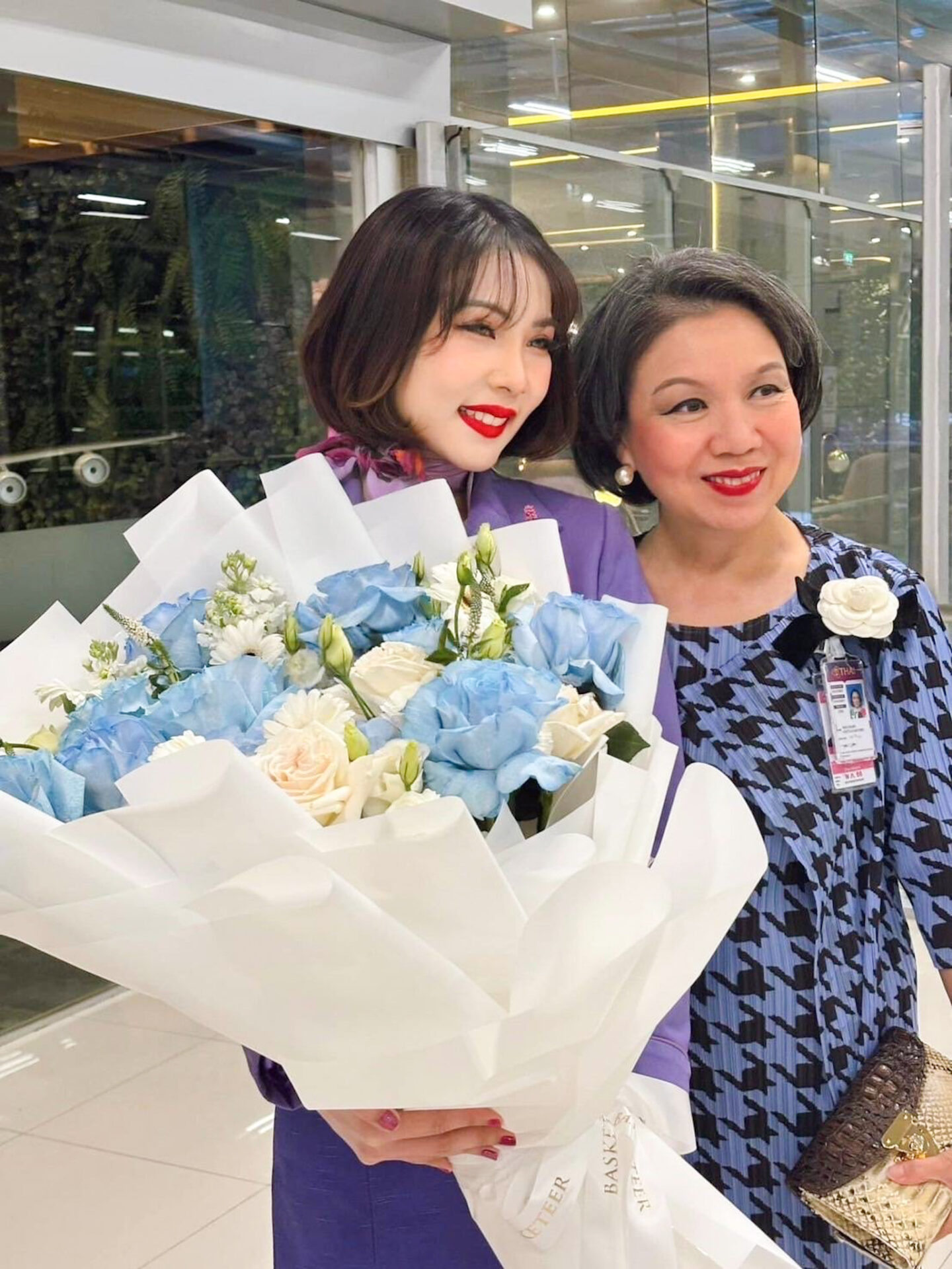 Two women, one holding a large bouquet of Serene Blue Bliss flowers, stand close together and smile. Both are dressed in elegant outfits, with the woman on the right also wearing a name badge and a flower pin. The background appears to be an indoor event space.