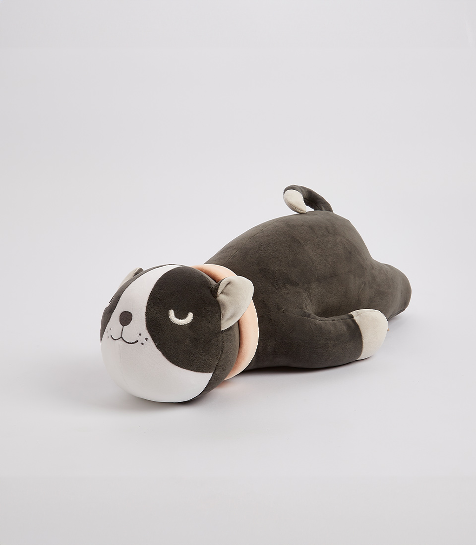 An adorable plush toy resembling a sleeping dog lies on its side against a plain white background. The soft toy features a dark brown body, light beige collar, and a face with closed eyes, a smiling mouth, and a white patch over one eye. Its legs and tail are stitched on, perfect for any collection of soft toys.