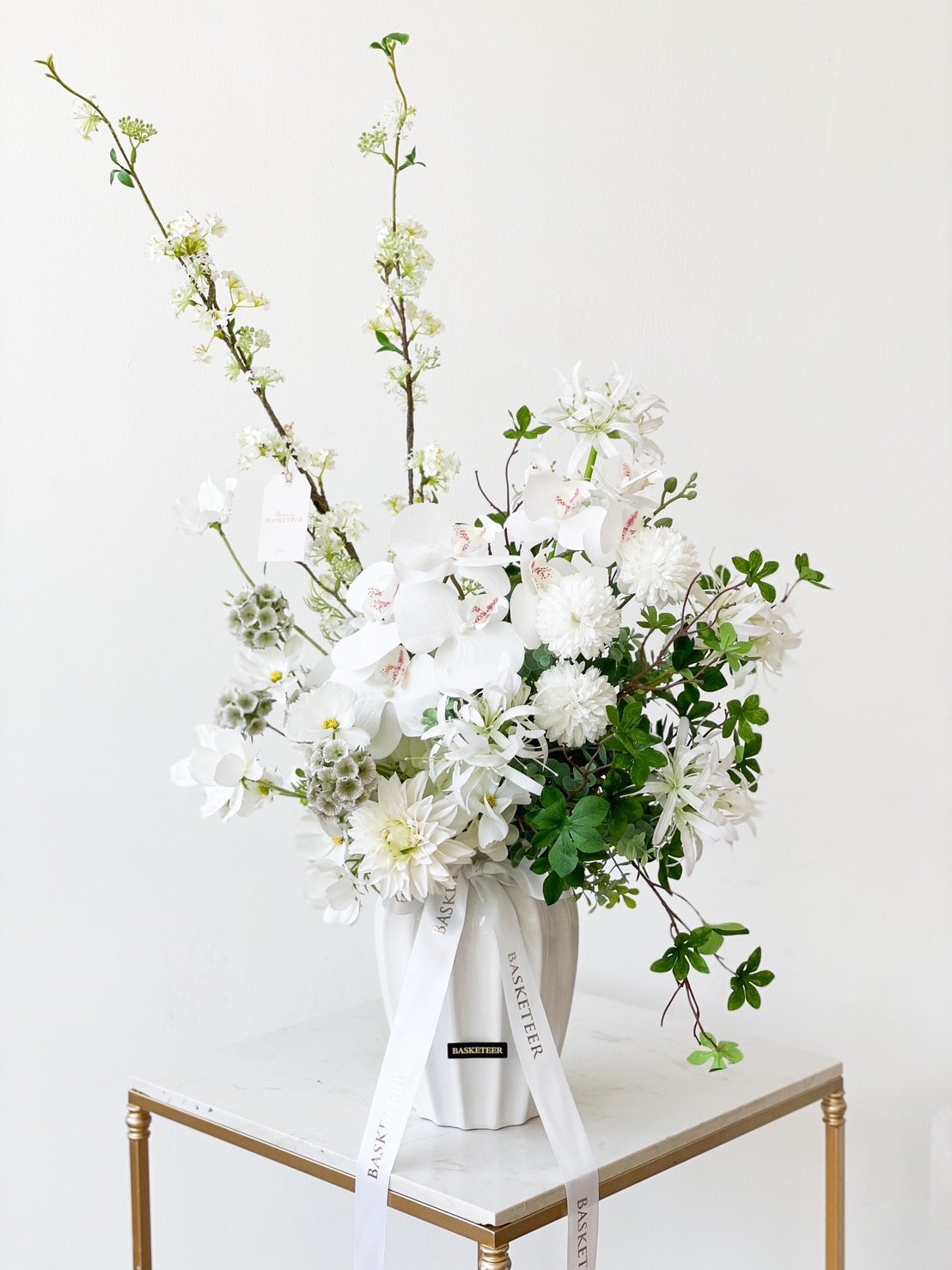 A frosty white garden artificial flower vase holds an arrangement of white and green flowers, including orchids, chrysanthemums, and leafy branches. The vase sits elegantly on a small white table with a gold trim, accessorized with white ribbons printed with text.