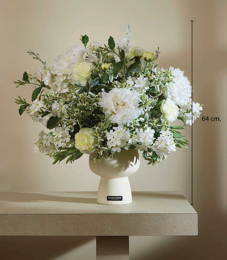 Artificial white flower arrangement with green foliage in a modern white ceramic vase on a table, displayed on a beige background, featuring a height measurement of 64 cm.