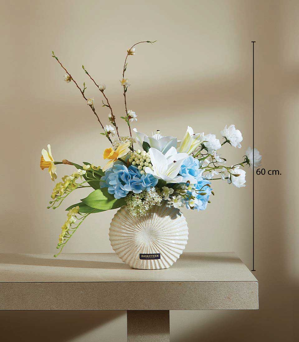 Artificial flower arrangement with white lilies, blue hydrangeas, and yellow daffodils in a white ceramic vase with black logo Basketeer, displayed on a beige background, featuring a height measurement of 60 cm.