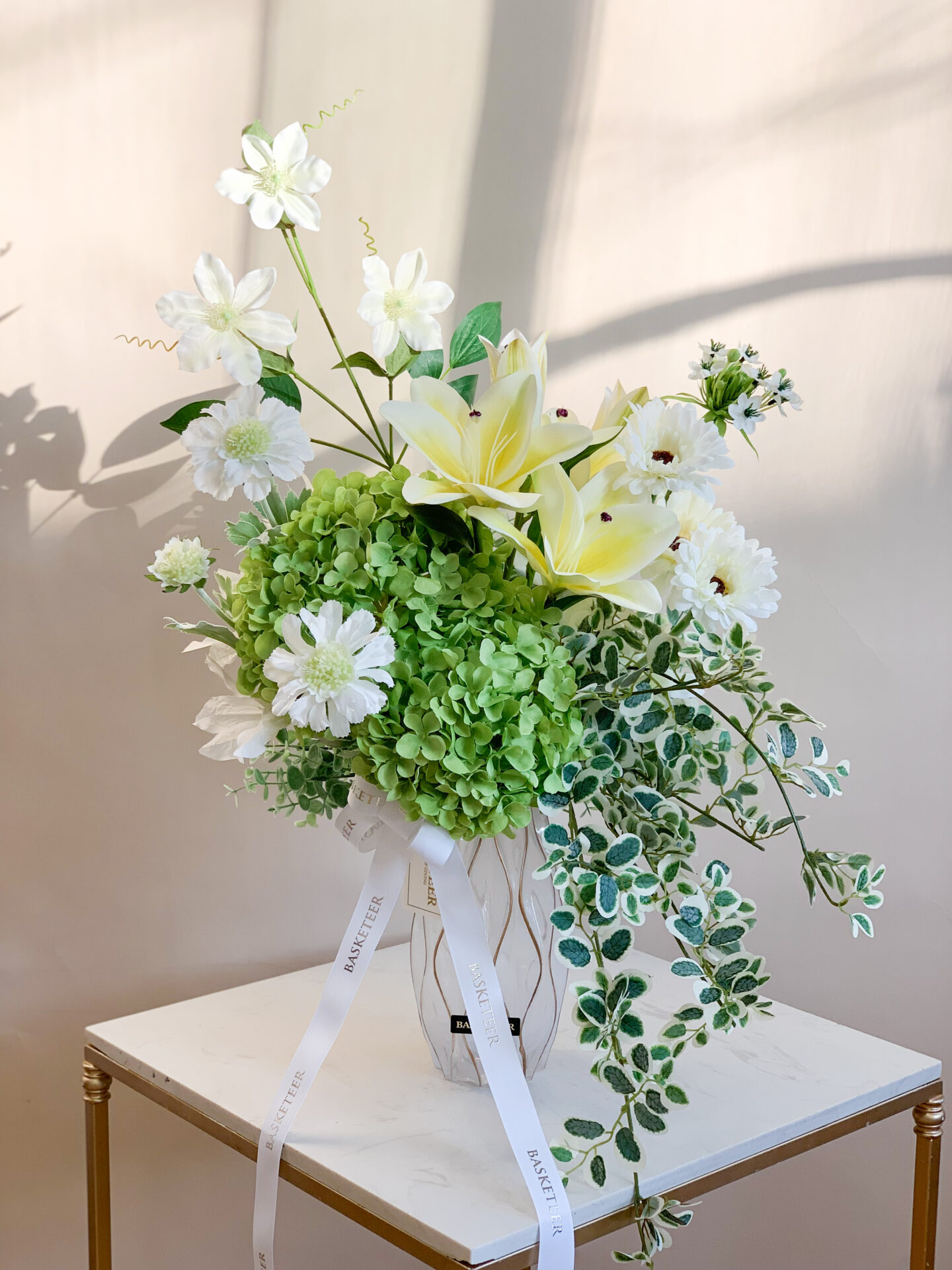 Artificial flower arrangement featuring white lilies, daisies, green hydrangeas, and foliage in a modern textured vase, displayed on a beige background.