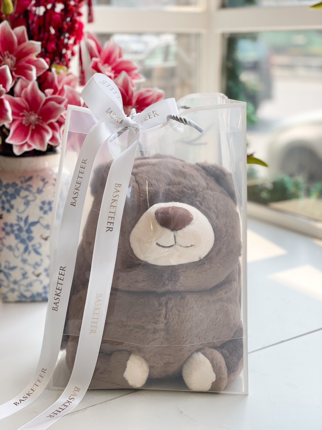 A soft and huggable brown bear plush toy in a transparent gift bag with a white ribbon.