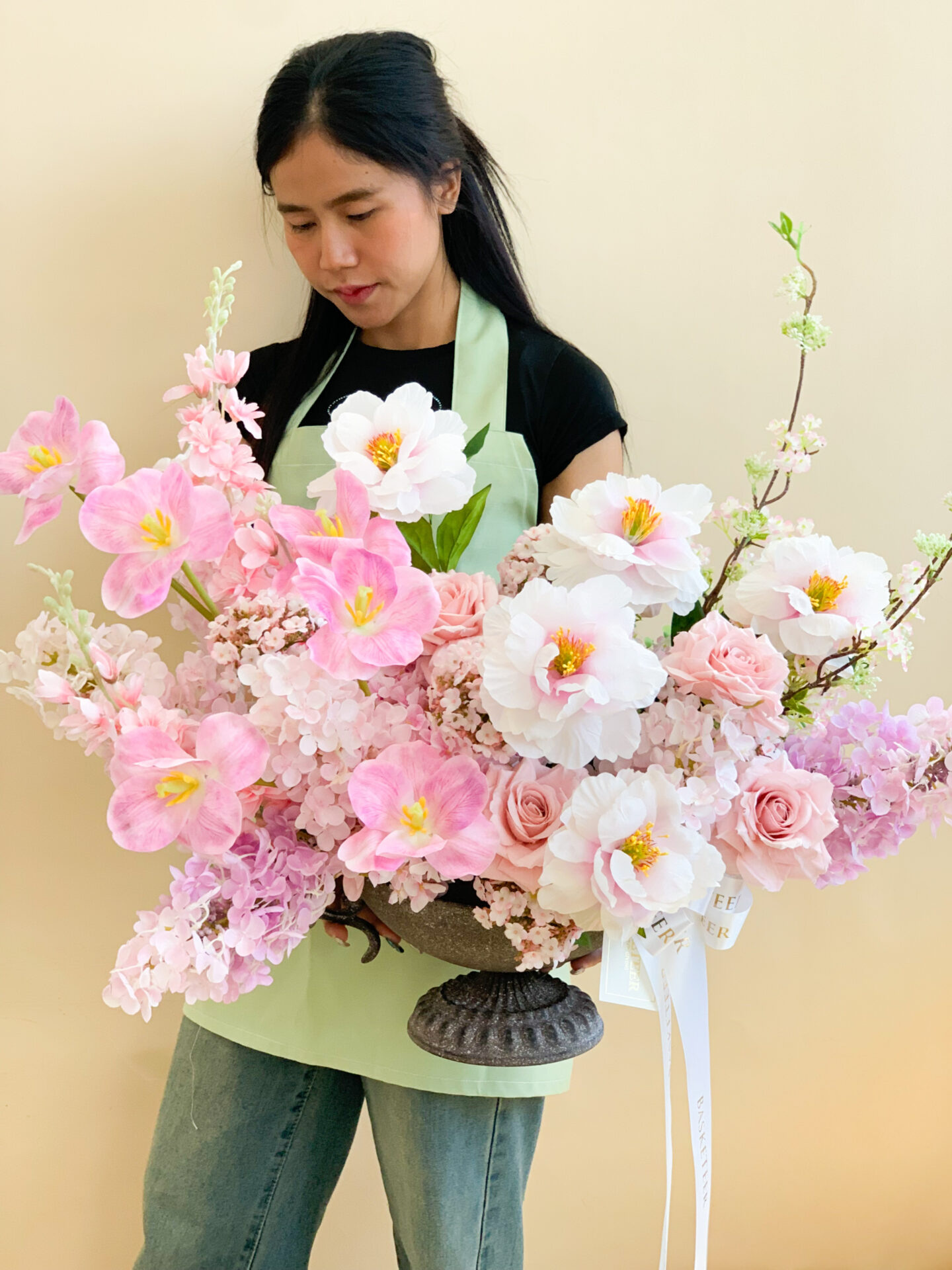 Artificial pink and white floral arrangement in an ornate pedestal vase on a beige table, displayed on a beige background.