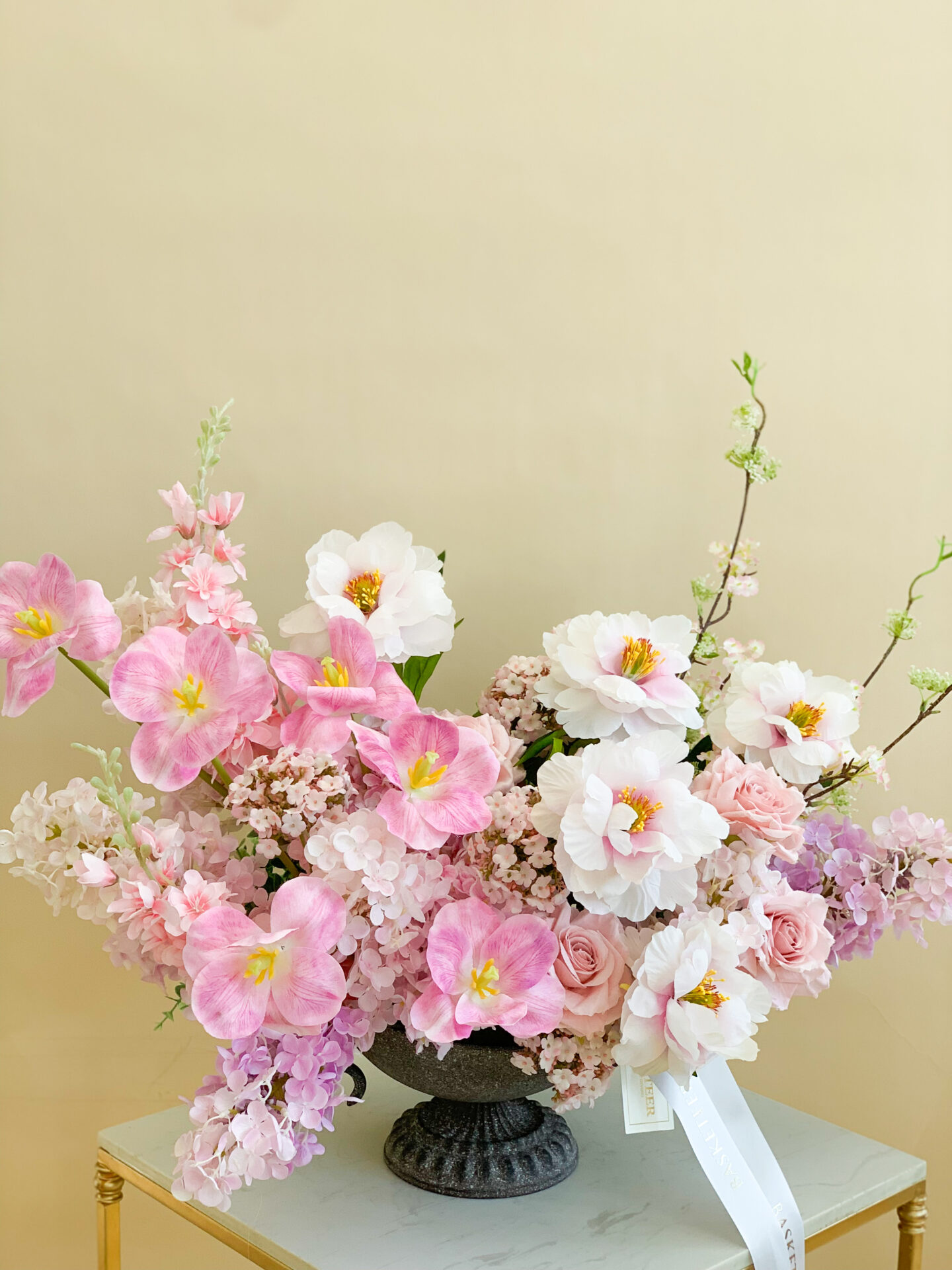 Artificial pink and white floral arrangement in an ornate pedestal vase on a beige table, displayed on a beige background.