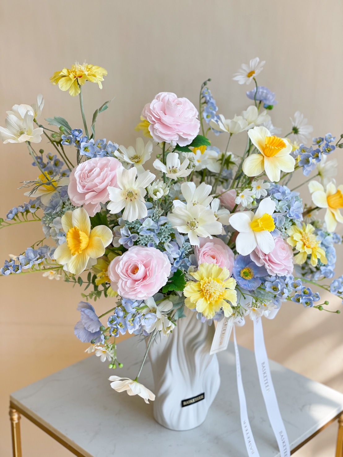 Pastel floral arrangement with pink roses, yellow daisies, white daffodils, and blue blossoms in a white sculptural vase.