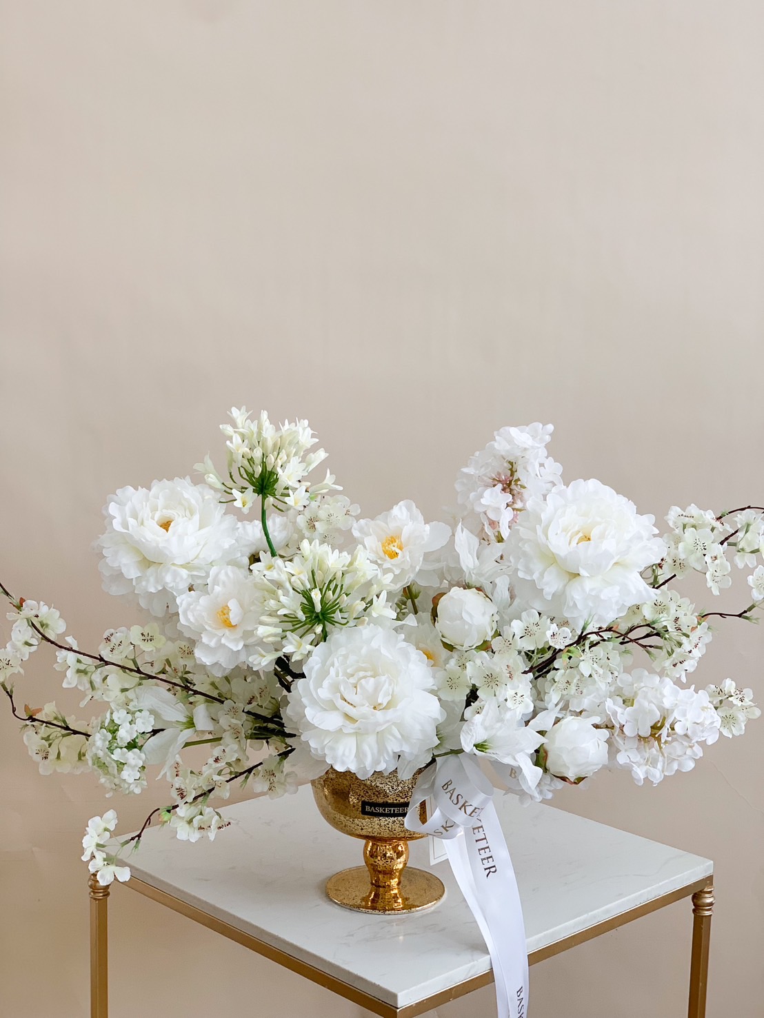 Elegant gold vase with a stunning arrangement of lifelike white flowers, including peonies and hydrangeas, on a beige background.