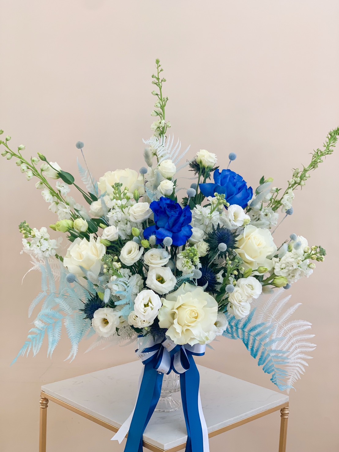 Elegant blue and white floral arrangement with roses and hydrangeas in a white vase, adorned with a blue ribbon.