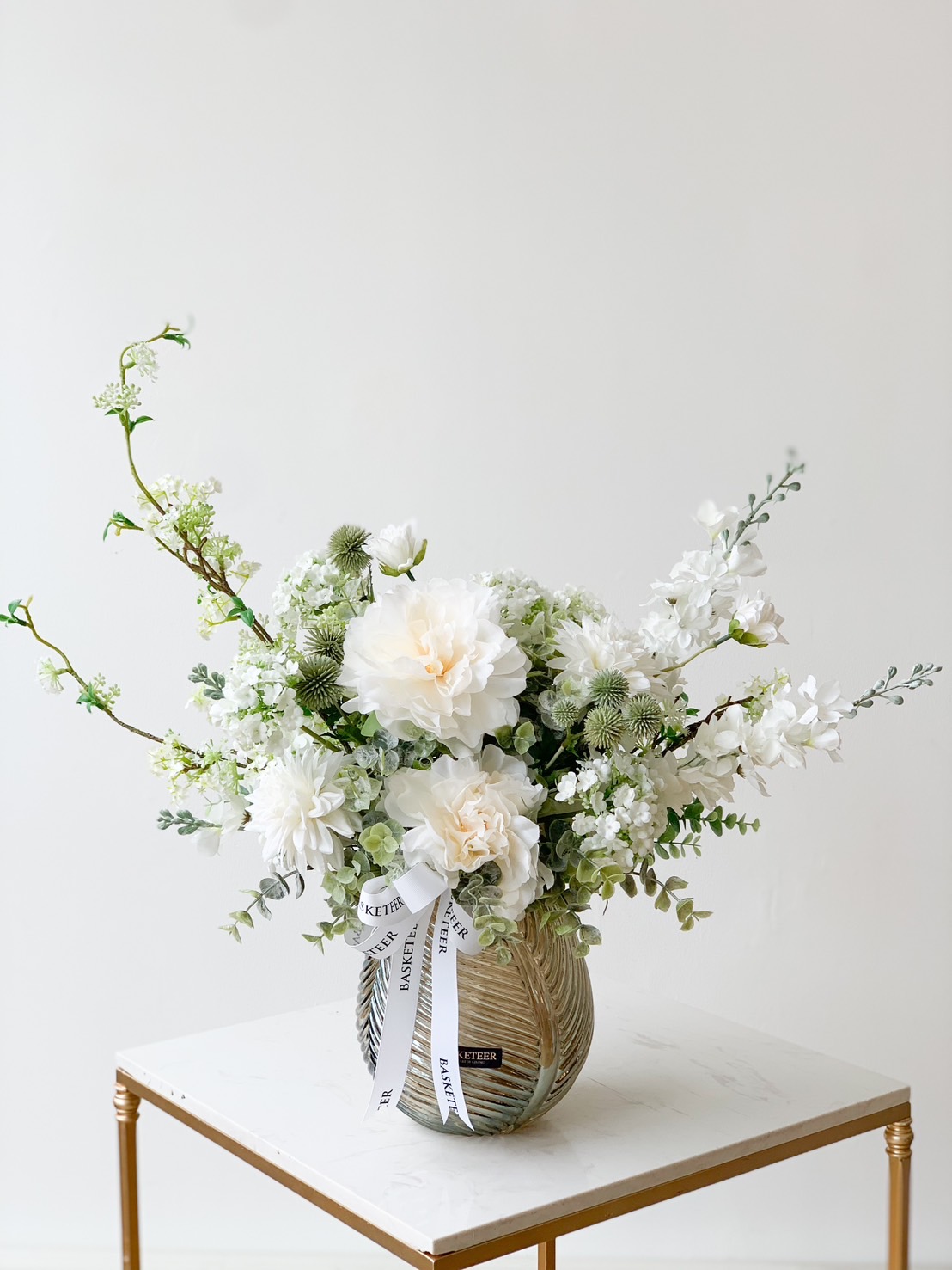 A lush and sophisticated arrangement of white and pale green blooms, featuring exquisite peonies, hydrangeas, and delicate fillers graces a textured white vase. The vase is elegantly adorned with a luxurious ribbon imprinted with 