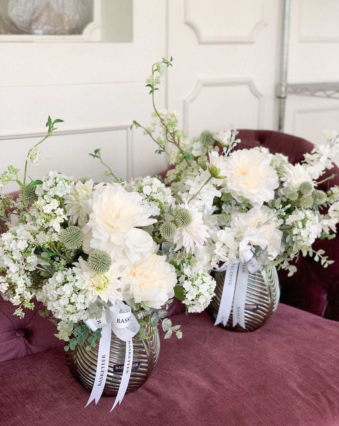 Two exquisite floral arrangements in opulent glass vases grace a sumptuous purple velvet bench. Each sophisticated bouquet showcases pristine white flowers, lush greenery, and elegant white satin ribbons adorned with the words 