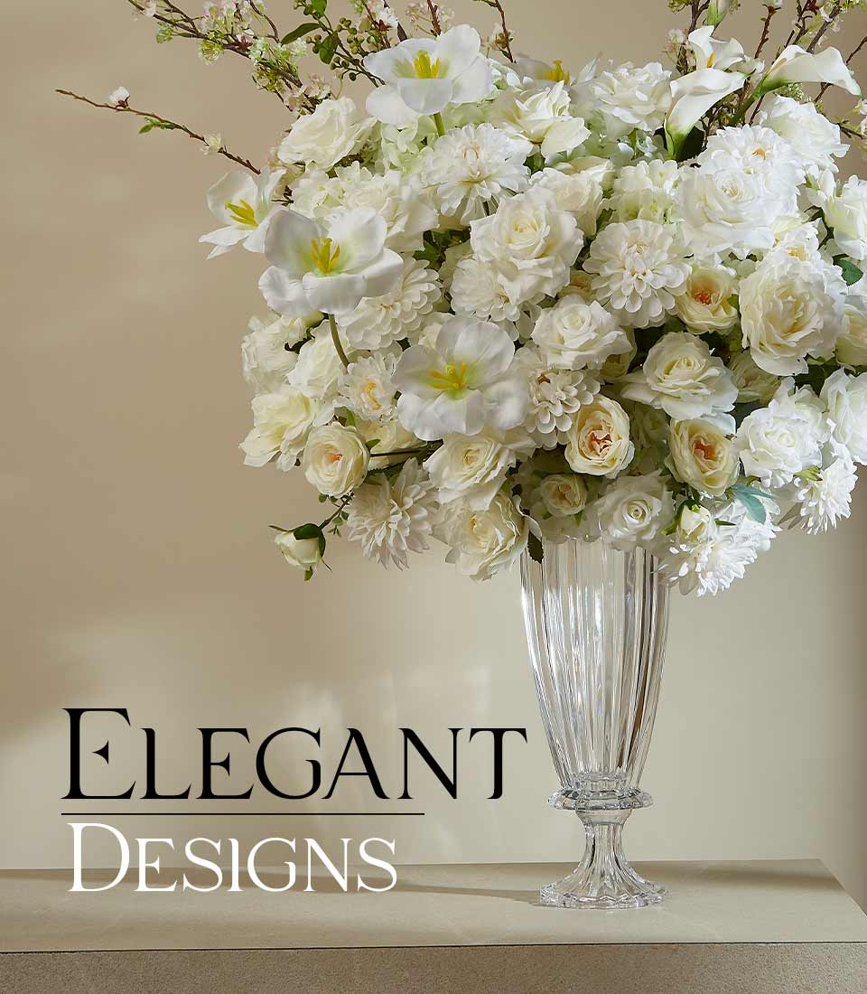 A towering clear glass vase cradles a sumptuous bouquet of lifelike artificial white flowers, featuring pristine roses and opulent dahlias. Set against a backdrop of soft beige, this exquisite arrangement embodies pure elegance and serenity. A sophisticated ribbon adorned with the Basketeer logo embellishes the vase, while the text 