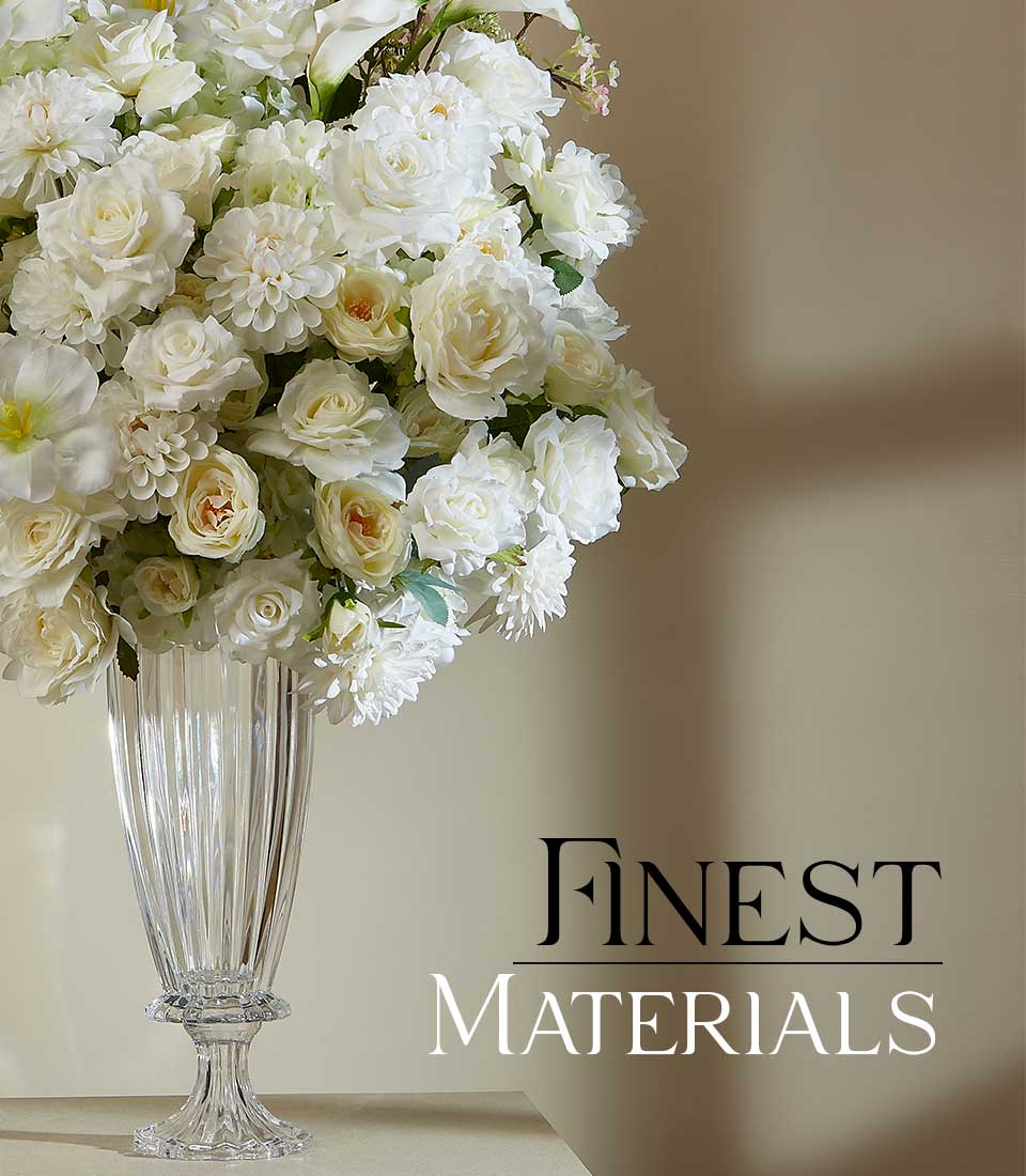A crystal-clear glass vase, holding an opulent bouquet of pristine white roses and dahlias, radiates elegance against a minimalist backdrop. The luxurious floral arrangement epitomizes the sophisticated allure of Basketeer. A ribbon with the Basketeer logo subtly complements the composition, enhancing its refined presentation. To the right, 