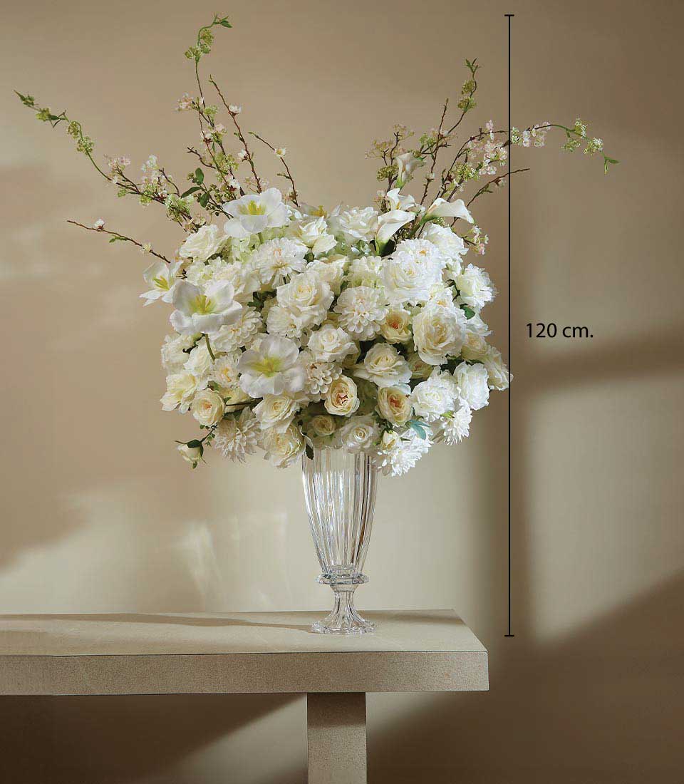 A luxurious arrangement of pristine white artificial flowers, standing majestically at 120 cm, graces a refined beige table against a backdrop of an elegantly light brown wall. This exquisite floral display, housed in a clear glass vase, boasts stunning roses and lilies interwoven with elegant green branches. The bouquet epitomizes timeless beauty and is complemented by the distinctive Basketeer logo on an accompanying ribbon or box, accentuating its sophisticated appeal.
